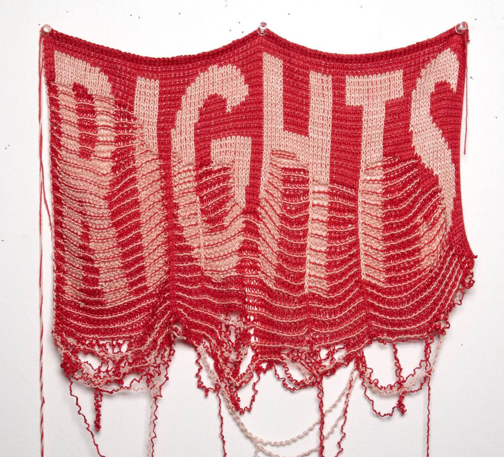 A knitted red banner with big pink letters that say “Rights.” The banner unravels towards the lower part, making the word “Rights” disappear.