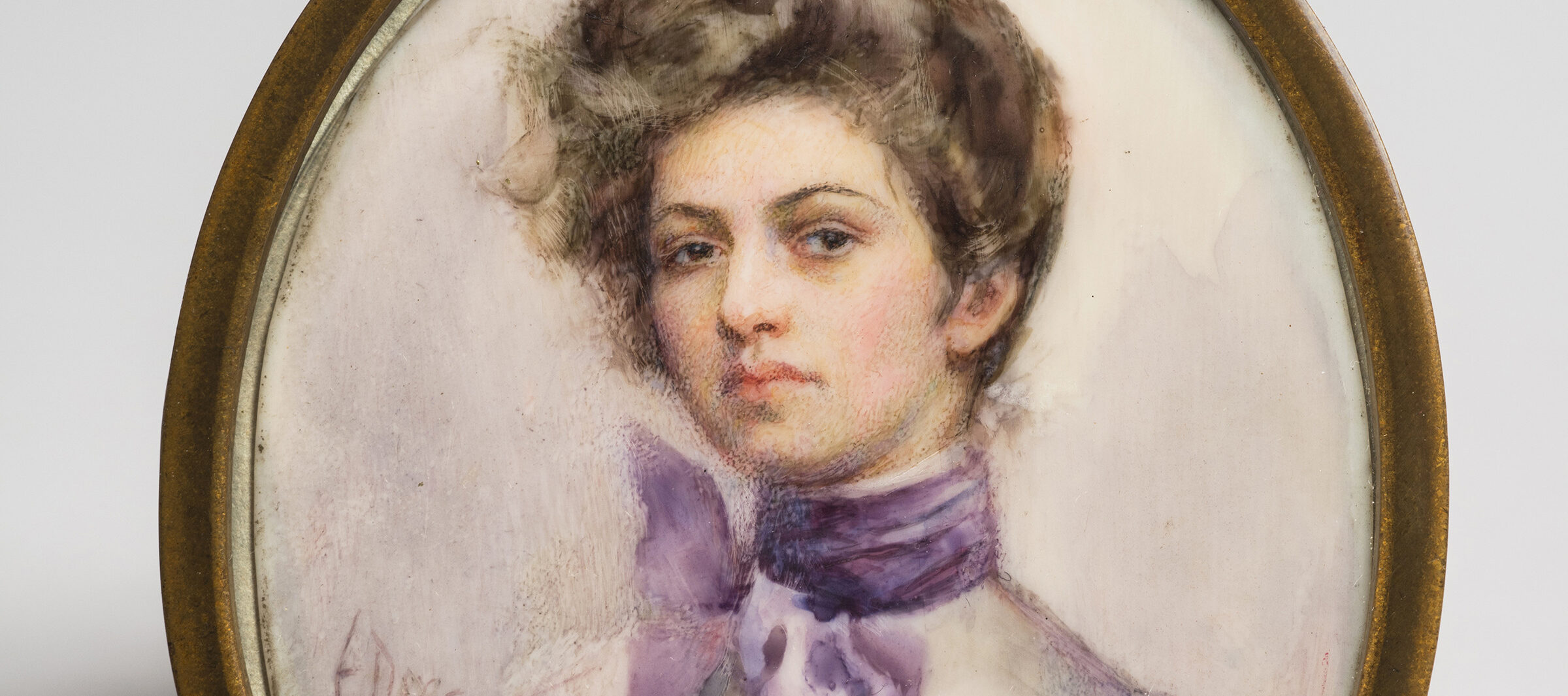 Miniature half-portrait of a light-skinned woman, set in an oval-shaped brass frame, gazing confidently at the viewer.  The woman is wearing a Victorian-style white blouse with a purple scarf tied in bow around her neck, her brown hair loosely piled atop her head.