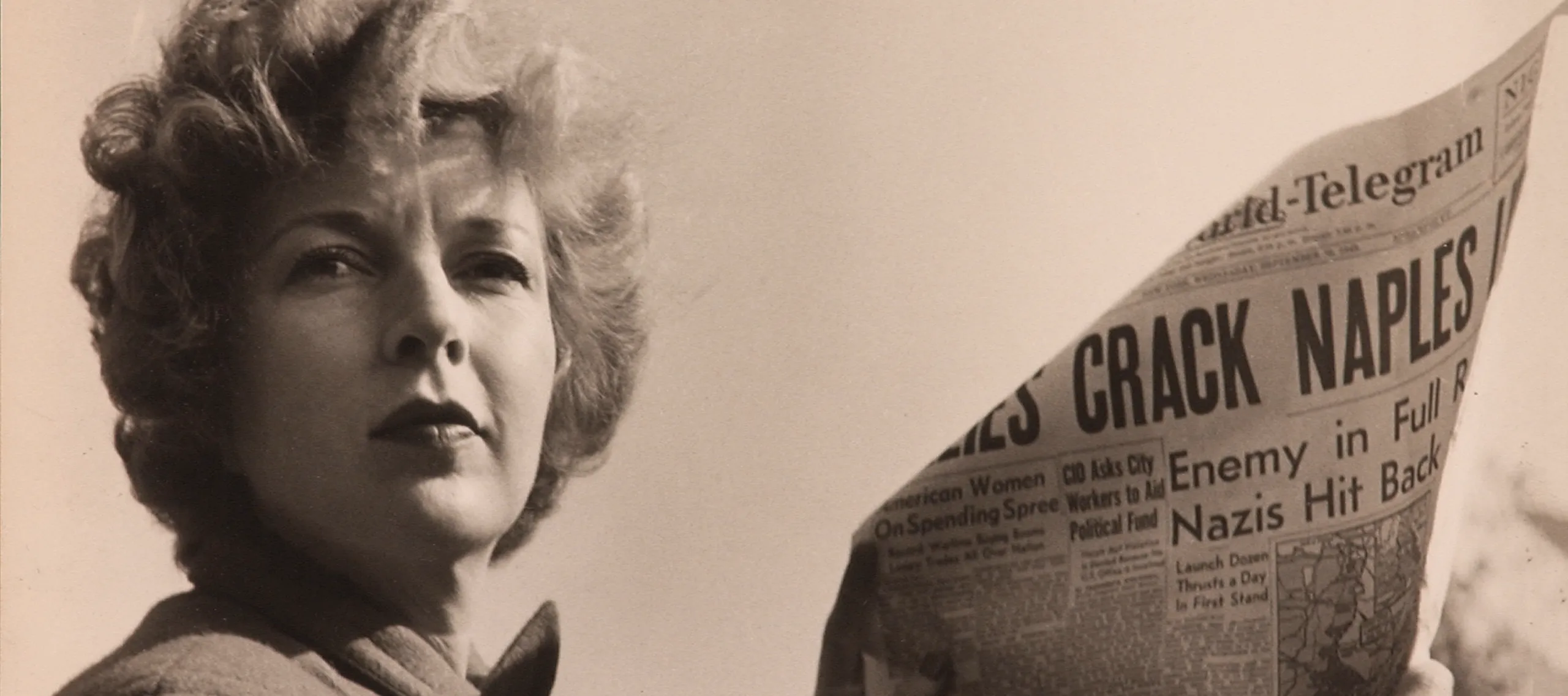 A black-and-white photograph of a light-skinned adult woman holding a newspaper with news about World War II. She wears a coat and her short, curly hair is caught in the wind.