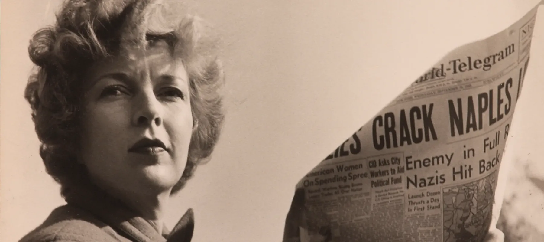 A black-and-white photograph of a light-skinned adult woman holding a newspaper with news about World War II. She wears a coat and her short, curly hair is caught in the wind.