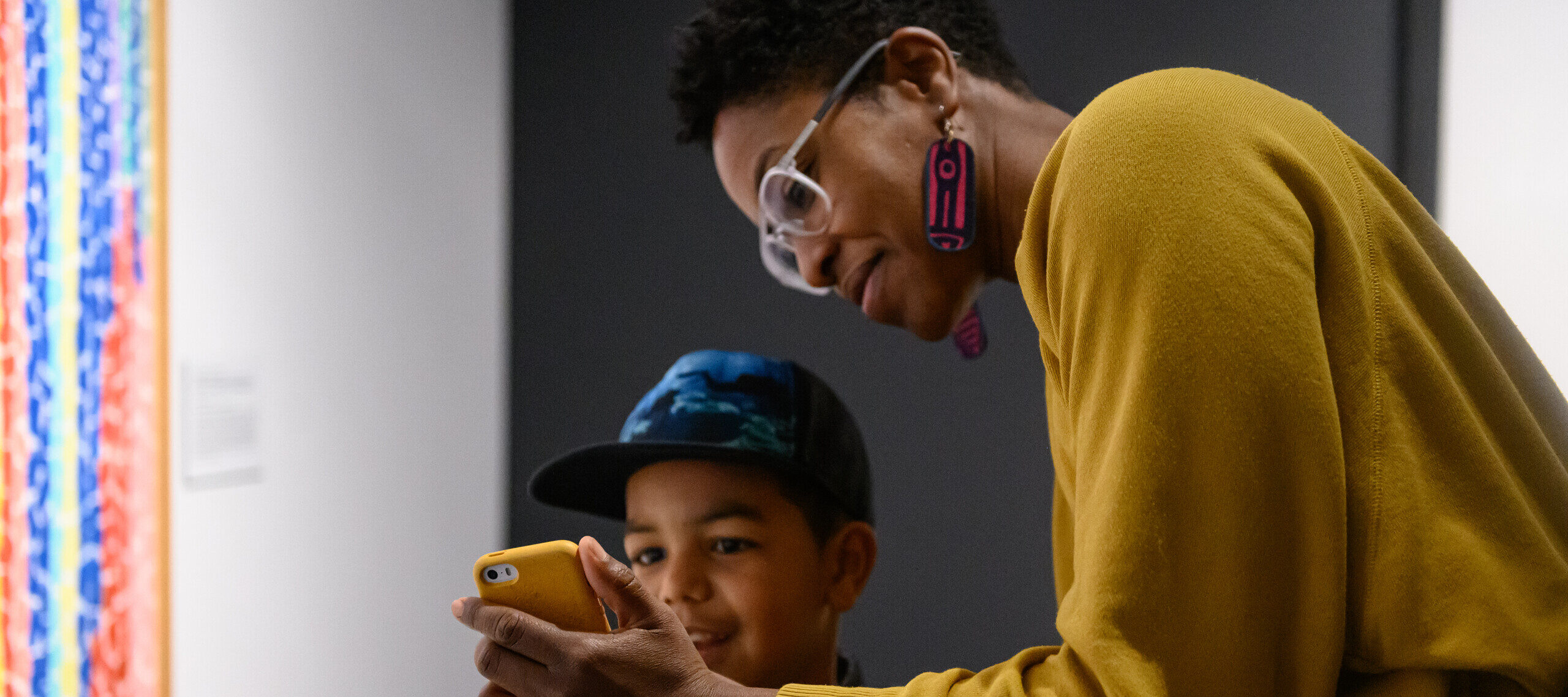 A dark-skinned woman wearing glasses shows a child something on the screen of her phone. In the background is a colorful work of art.