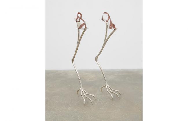 Two wiry metal objects stand on concrete before a white wall. The objects are in the shape of bird legs, with women’s shoes placed on top of them, which makes the bird legs look like stilts. 