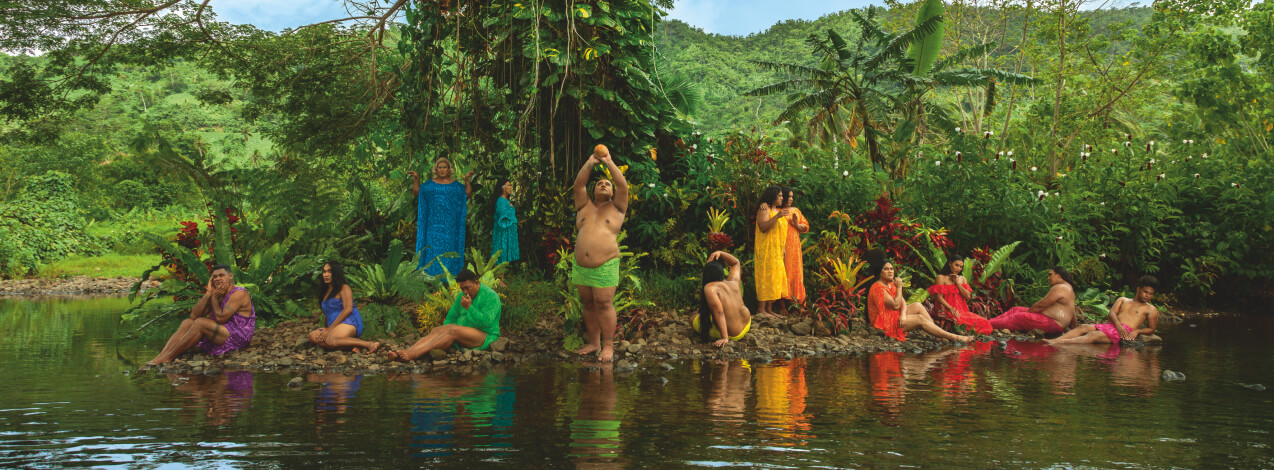 A photograph of several people with a medium skin tone sitting by a beach in front of a rain forest. The photograph is a staged scene, with the people posing in a way that is reminiscent of paintings.