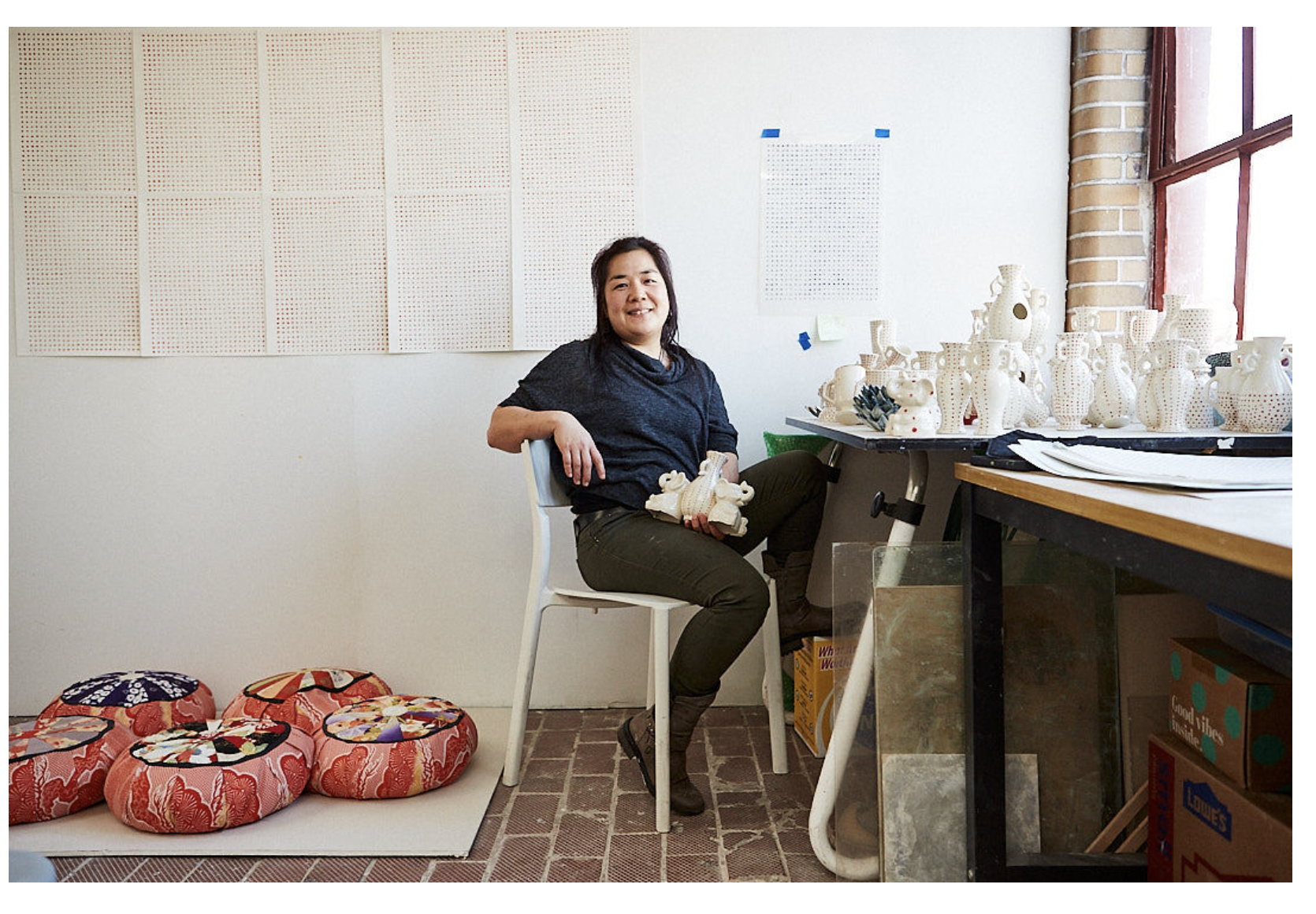 The artist sits in her studio, leaning back on a chair and holding one of her sculptures. She has long, black hair and a light skin tone, and she wears a blue sweater and olive green pants. The studio is very bright, with a white wall, several pink and red pillows scattered on the floor, and a whole table full of white vases. 