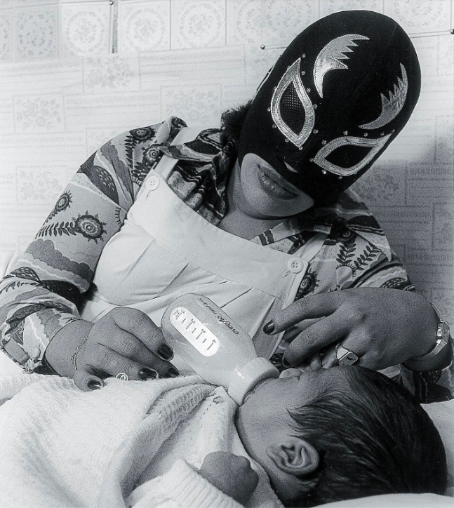 A black-and-white photograph of a woman wearing a wresting mask who is bottle-feeding a baby. The mask is black and gold and masks the woman’s face except for her mouth. She is wearing nail polish, lipstick, and a floral shirt that contrasts the wrestling mask traditionally associated with male wrestlers.