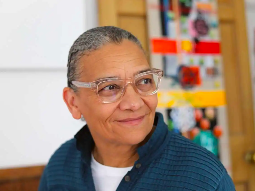 A close-up photograph shows a woman with a medium skin tone, glasses, and greyish black hair pulled back, who smiles and gazes into the distance. She is standing in front of a colorful piece of art, which is out of focus. 