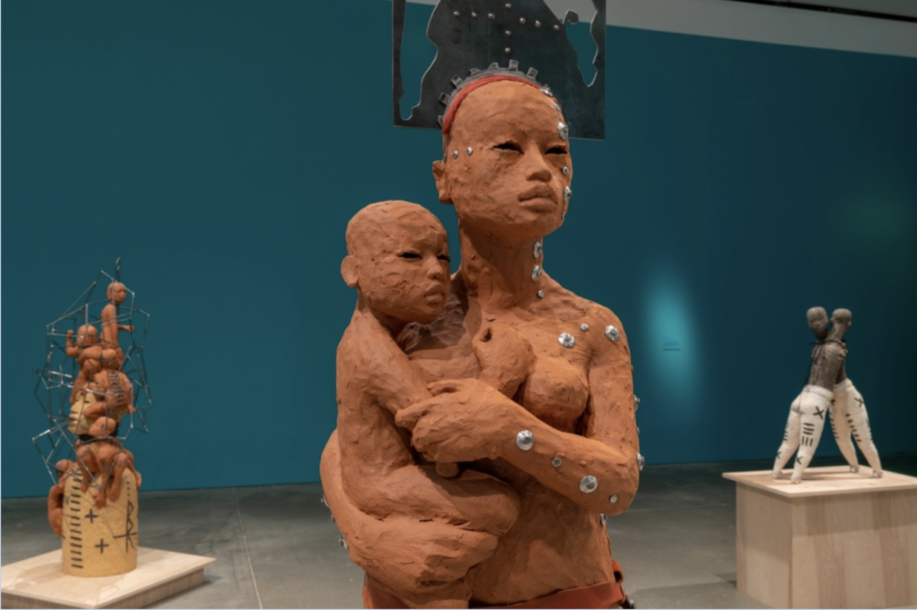An installation view of several ceramic sculptures in a room with a blue wall. The figure in the foreground is a woman holding a baby. The two figures are made of red clay, have a rough unfinished texture, and are bedazzled with round metal pieces.