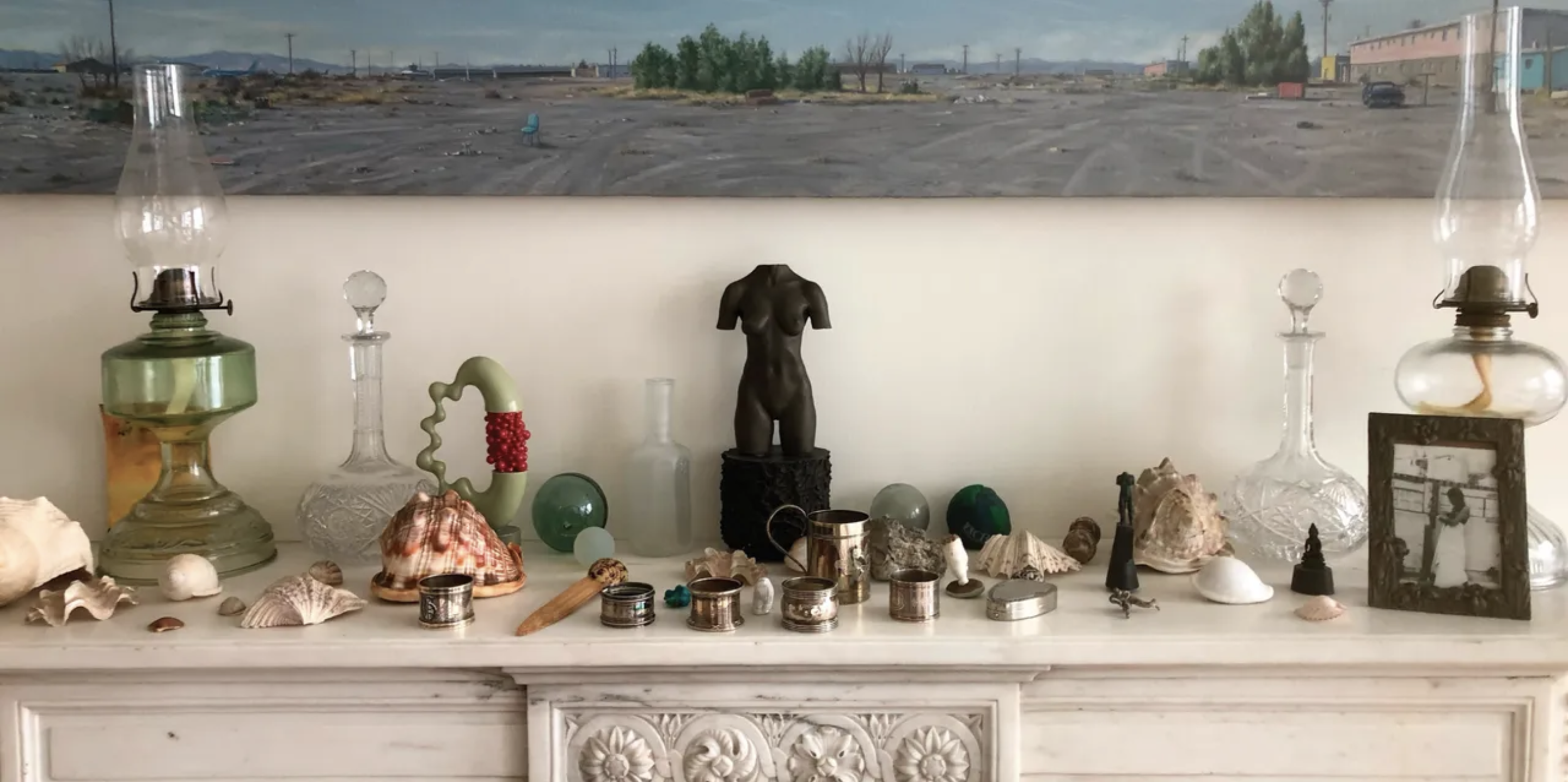 Several objects are placed on a marble fireplace: A small sculpture of a nude woman’s torso, seashells, lamps, metal cups, and glass carafes. A big painting depicting a landscape of a deserted parking lot with wide blue skies hangs above the fireplace.