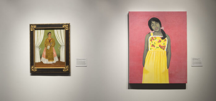 Two paintings are hanging on a white wall. On the left is a self-portrait of artist Frida Kahlo. She is wearing a pink dress and a yellow shawl, and is framed by two white curtains. The painting on the right is a portrait of a young girl with a gray skin tone wearing a yellow dress with strawberries.