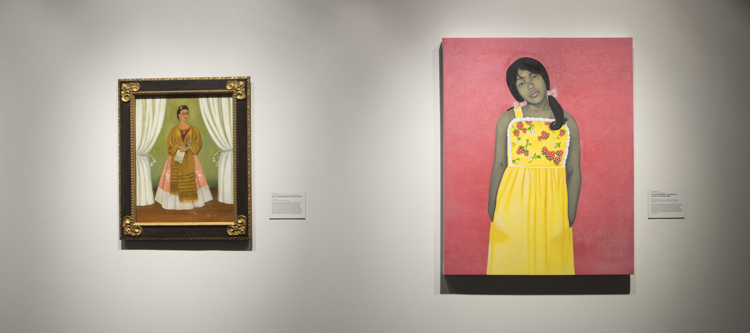 Two paintings are hanging on a white wall. On the left is a self-portrait of artist Frida Kahlo. She is wearing a pink dress and a yellow shawl, and is framed by two white curtains. The painting on the right is a portrait of a young girl with a gray skin tone wearing a yellow dress with strawberries.