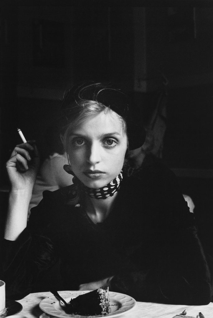 A black-and-white photograph of a young woman with a light skin tone and blonde hair, dressed in dark colors. The woman is sitting at a table smoking, and in front of her is a piece of cake. Her unflinching gaze is fixed on the viewer. 