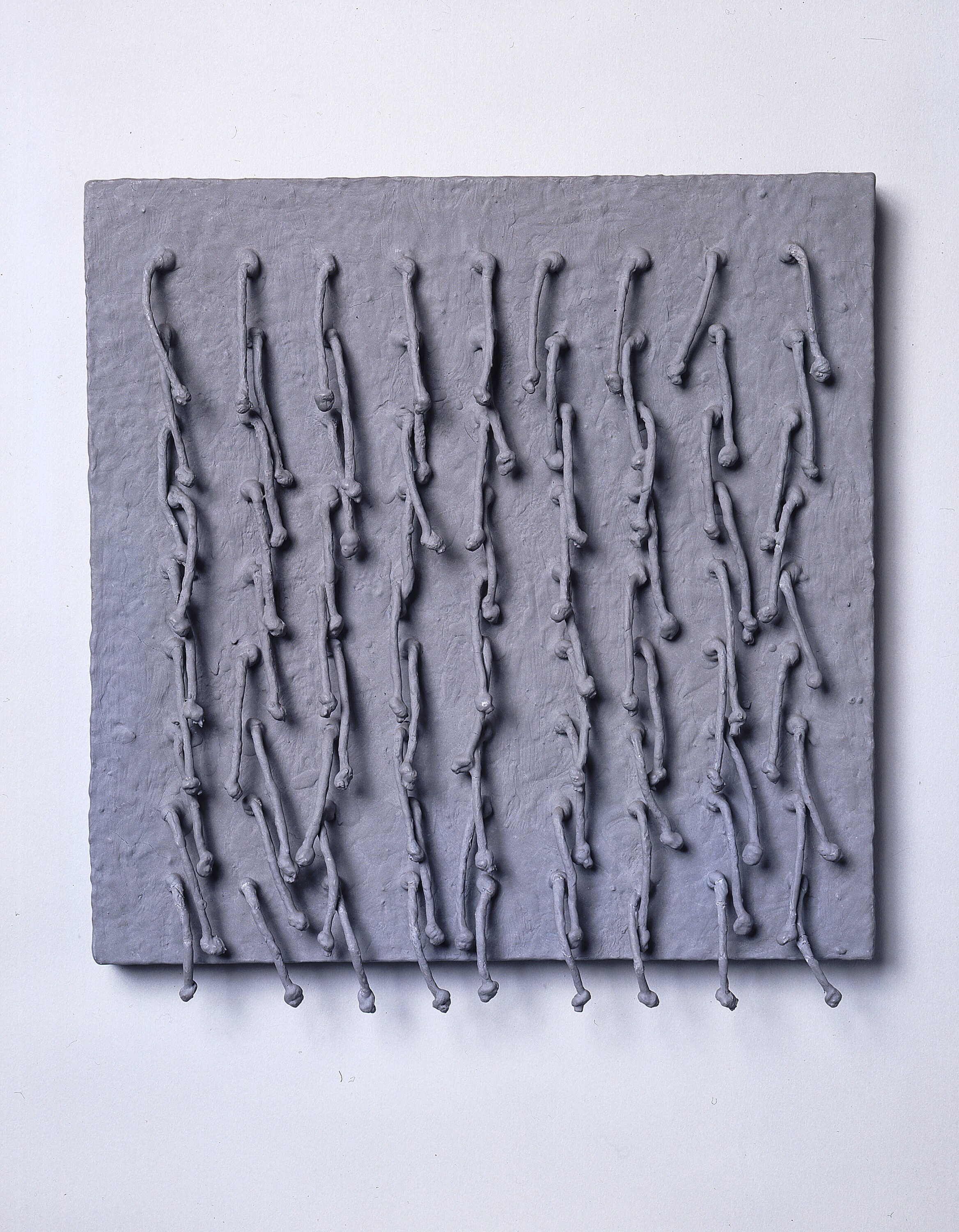 Short, knotted cords, arranged in a symmetrical 9-by-9 grid, emerge from a square panel and hang down. Thick, matte paint in a silvery gray color coats the entire piece, adding rough texture to the panel and stiffening the cords into positions that cast irregular shadows across the work’s surface.