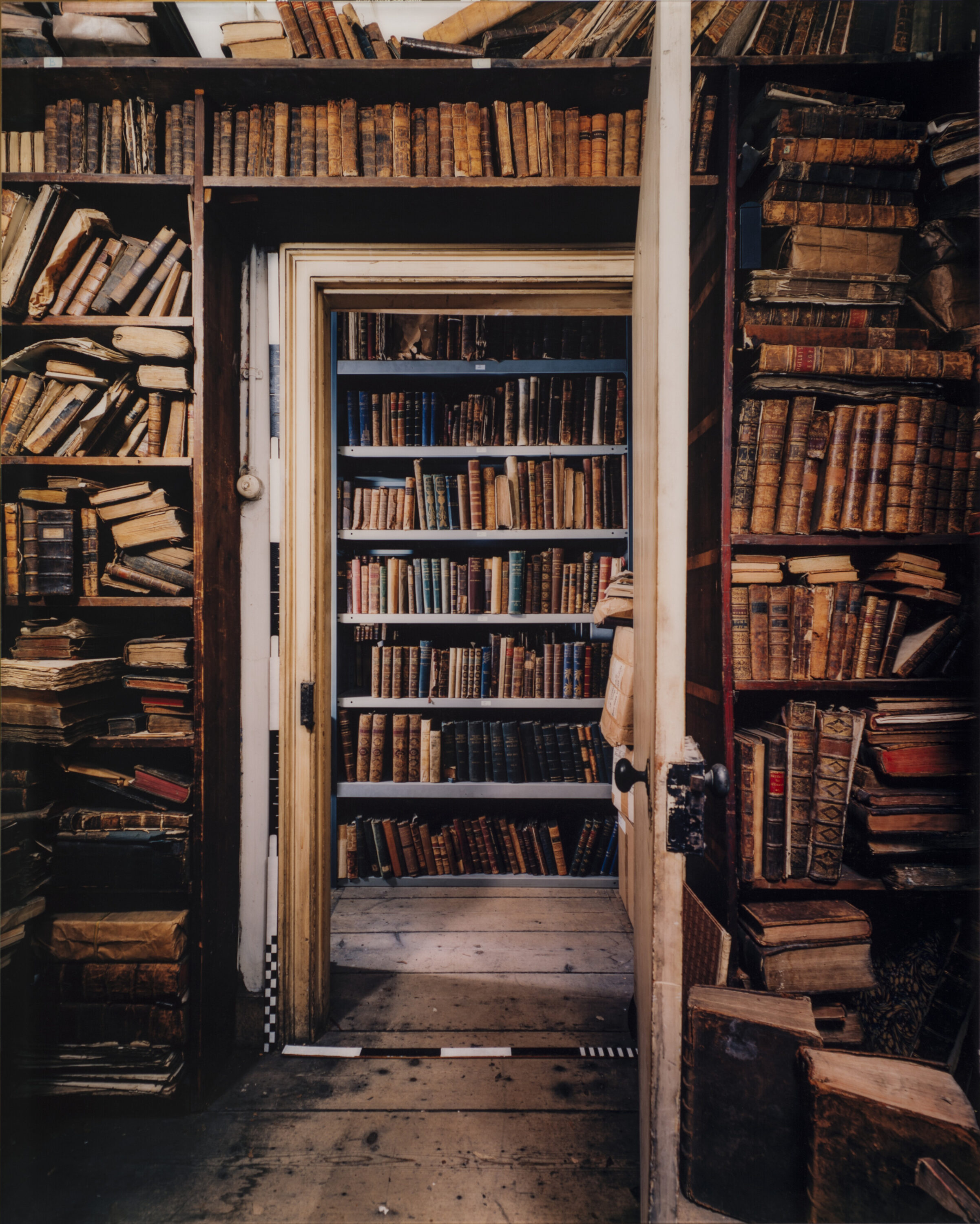 Color photograph of room with old books on bookshelves that stretch from floor to ceiling. In the center is an open door revealing another room with floor to ceiling shelves filled with books.