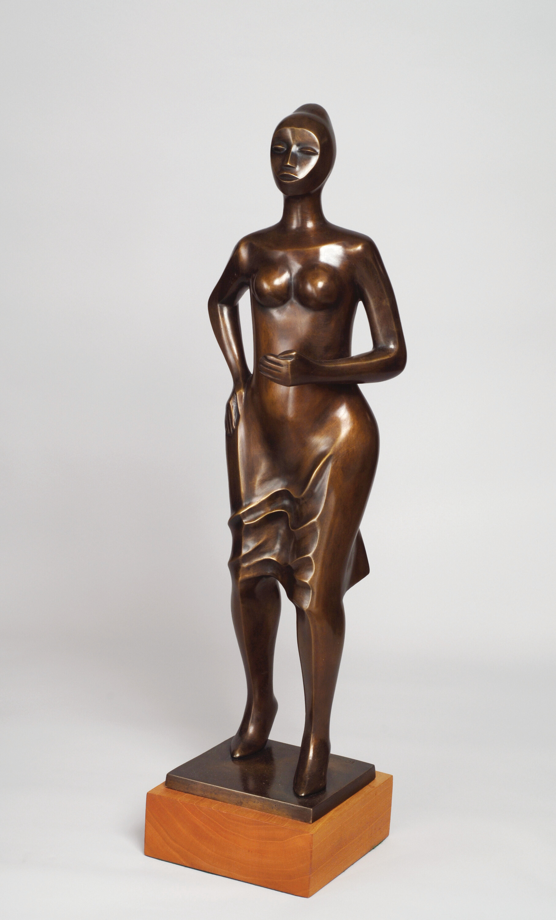 A bronze statue in warm brown shows the figure of a woman who gently steps forward, hand on hip. The forms of her body are stylized and geometric, and she wears a dress or skirt that is revealed where it gathers across her legs as if pulled by a breeze. Her face is simplified, upturned, and her hair is styled in a high bun. 