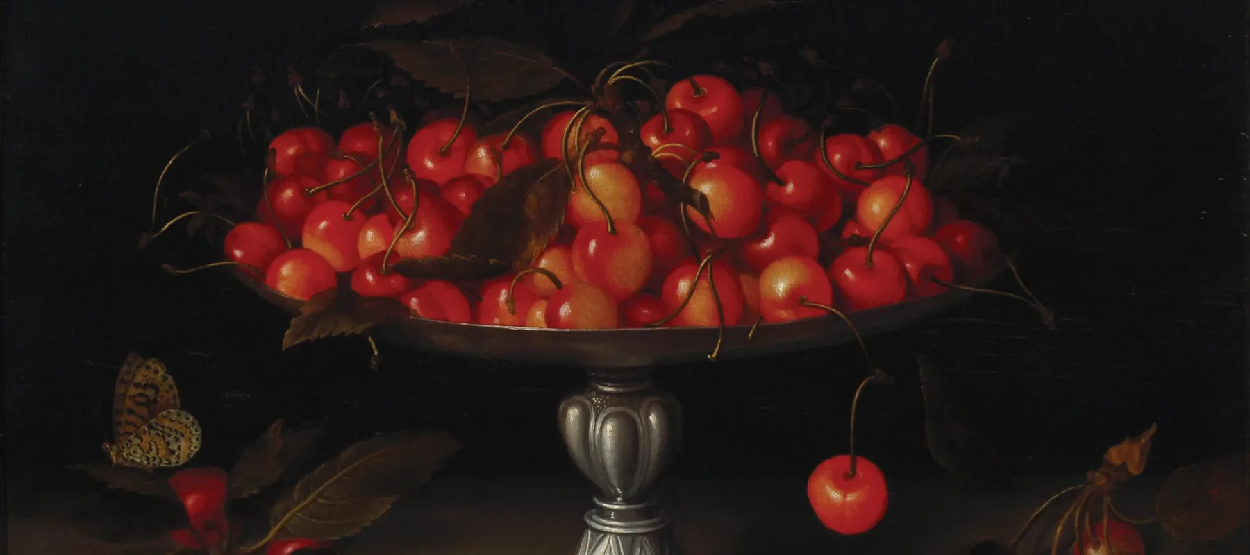 An opulent, footed silver bowl overflows with ripe cherries in front of a dramatic dark background. Additional fruits lie on the table below the bowl, and a butterfly flutters to one side.