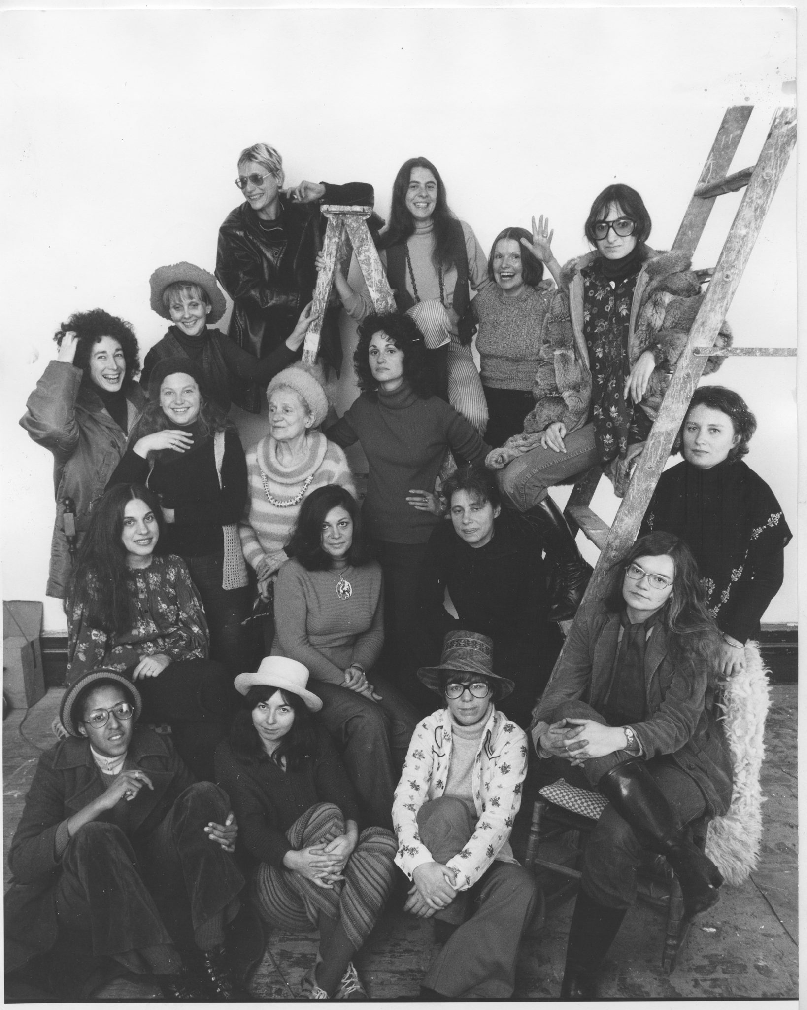 A black-and-white photograph shows a group of women from an elevated perspective. The women have light, medium, and medium-dark skin tones and smile at the camera. Many of them are wearing eccentric hats, beanies, and sunglasses.