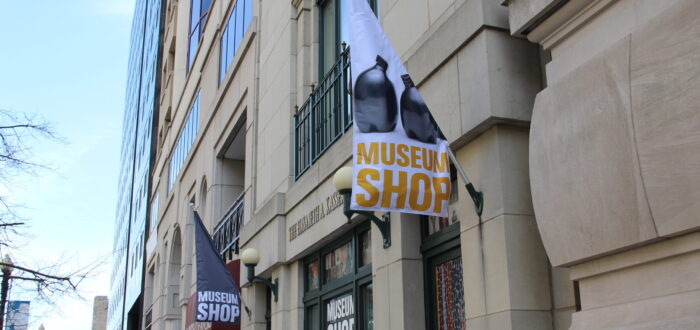 A white flag hangs on the facade of a stone building. The flag has an image of two black ceramic vases and "Museum Shop" written in gold letters below them.