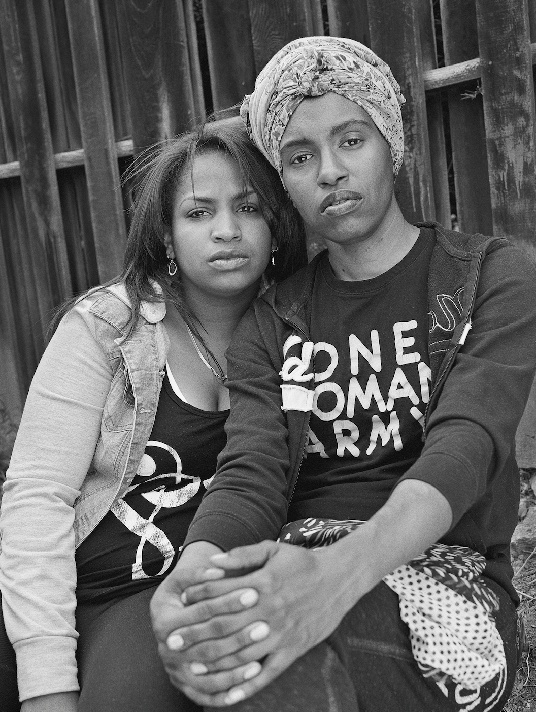 A black-and-white portrait of two women with a dark skin tone. The woman on the right wears a head wrap and a black T-shirt that says “One Woman Army” in white capital letters. The woman on the left is leaning on her shoulder. Both look into the camera with a skeptical expression on their face.