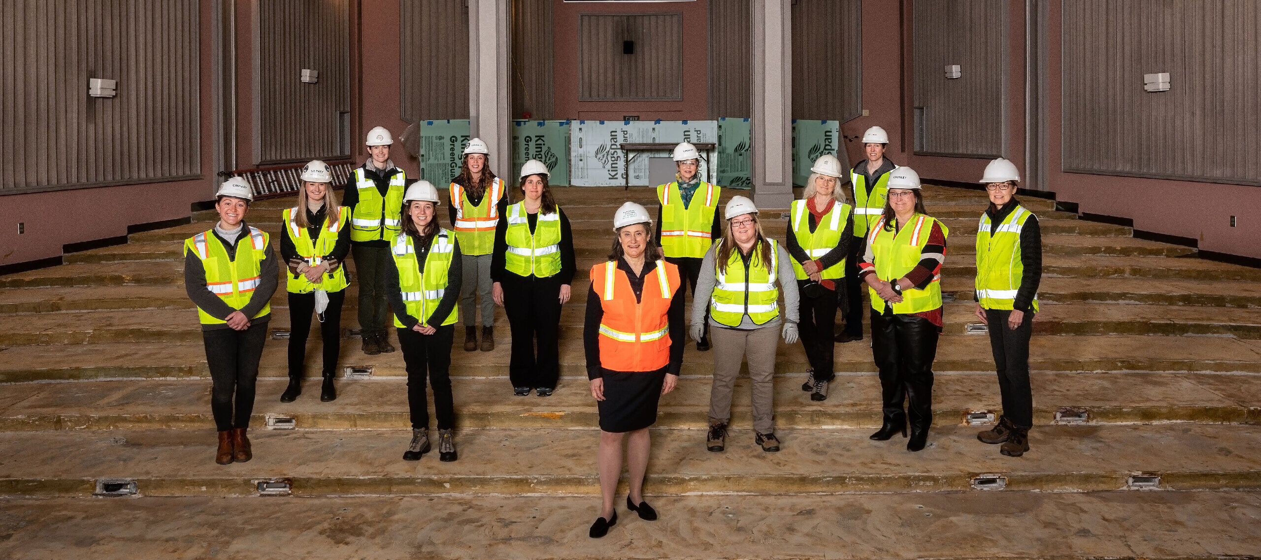 A group of thirteen women with a light skin tone of all ages wearing yellow safety vests and white safety helmets, smiling toward the camera. They are standing on a construction site with unfinished floors and building materials leaning on the walls in the back. One woman in the middle is wearing an orange safety vest, standing apart from the other women.