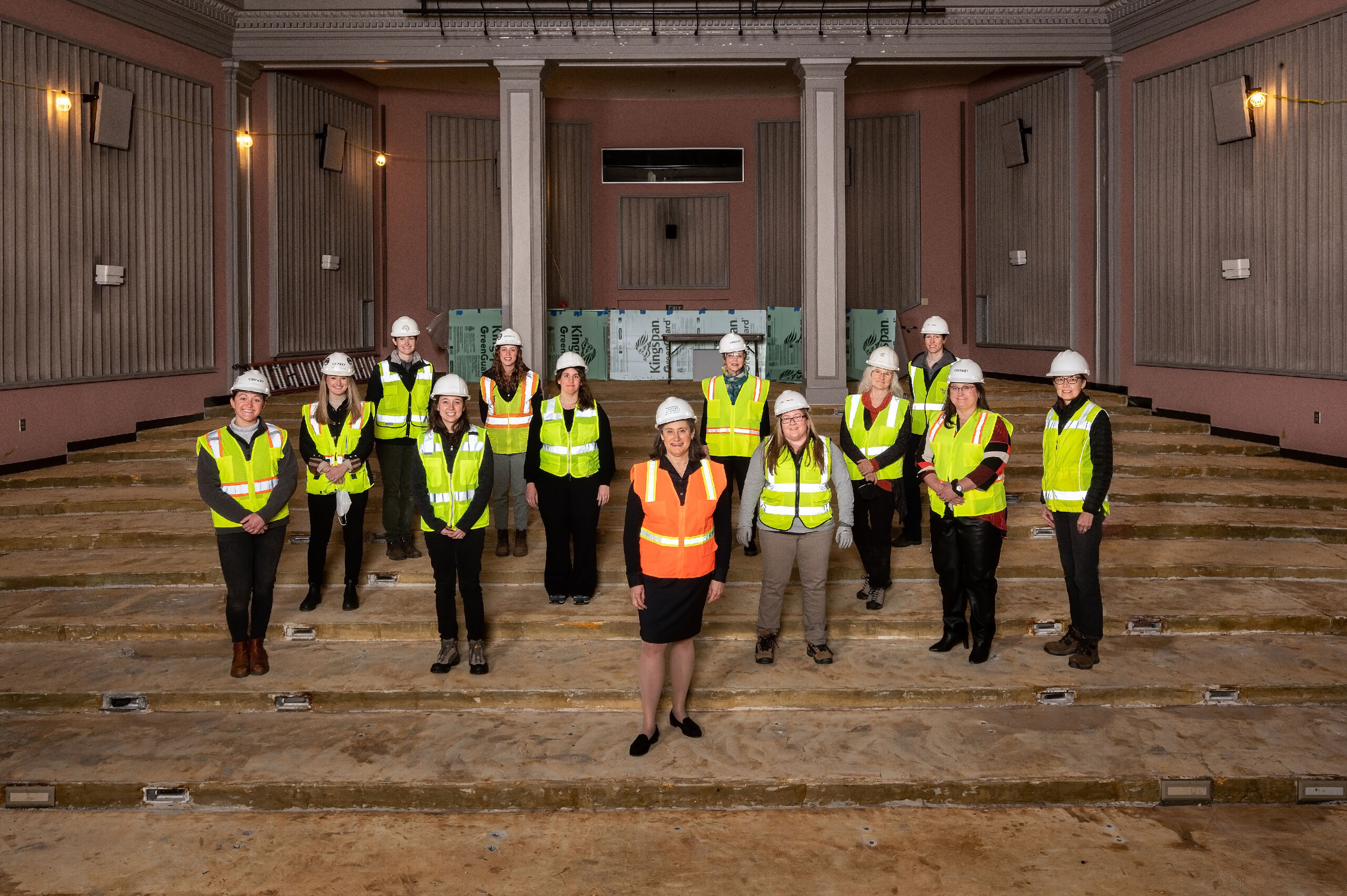 A group of thirteen women with a light skin tone of all ages wearing yellow safety vests and white safety helmets, smiling toward the camera. They are standing on a construction site with unfinished floors and building materials leaning on the walls in the back. One woman in the middle is wearing an orange safety vest, standing apart from the other women.