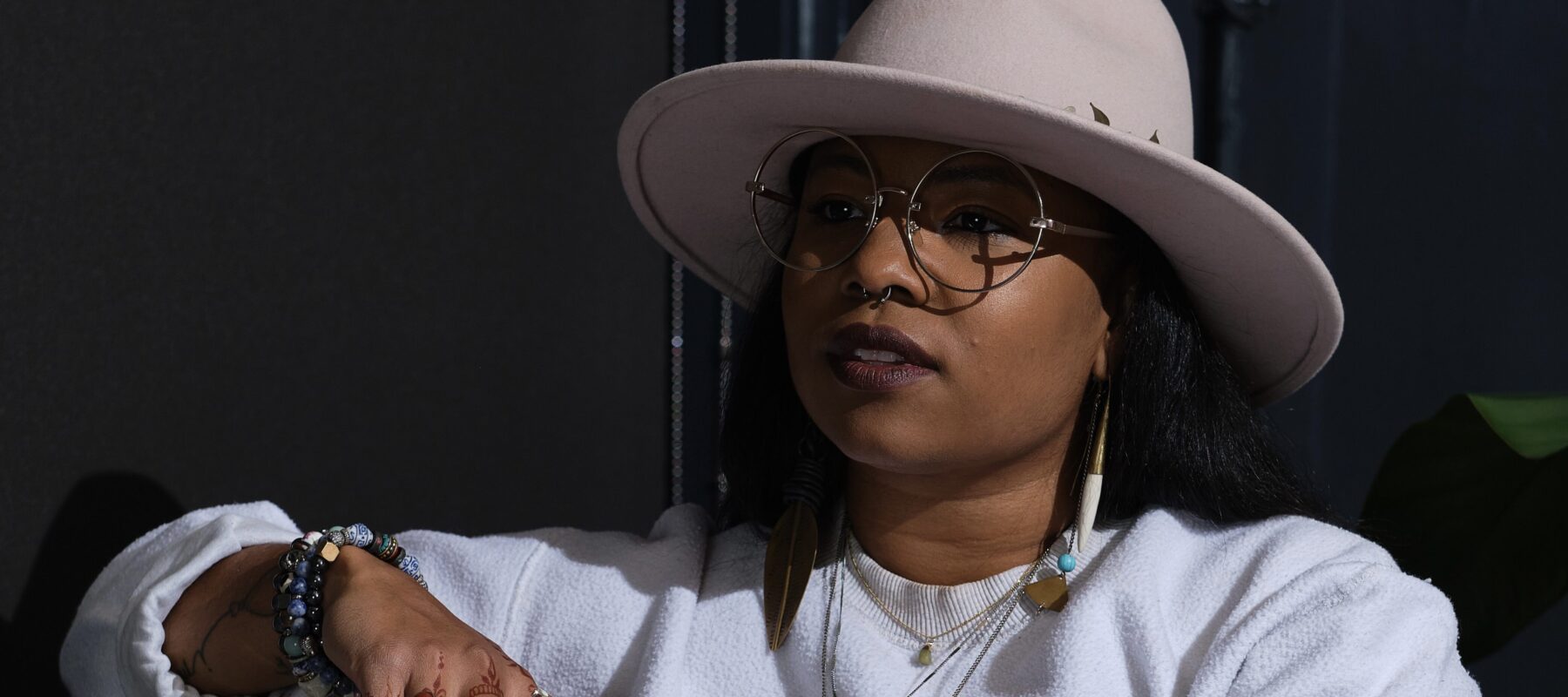 A medium-skinned woman with long brown hair and wire framed glasses sits on a couch in front of a charcoal wall. She is wearing a wool rancher hat, a long sleeved white sweatshirt, a series of long gold necklaces, several beaded bracelets, and has henna tattoos on her fingers.