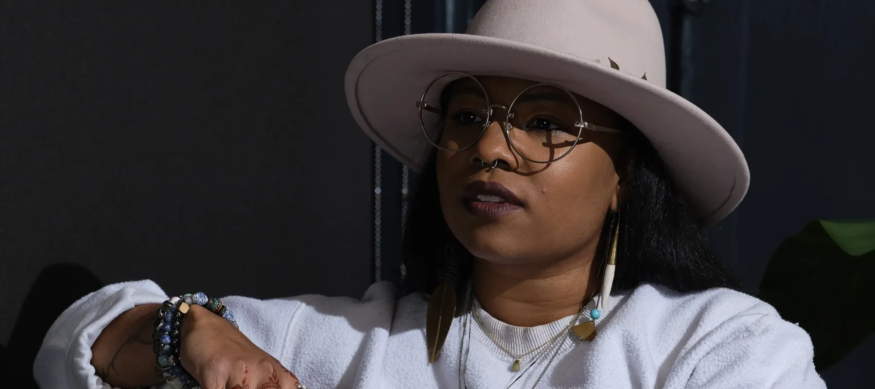 A medium-skinned woman with long brown hair and wire framed glasses sits on a couch in front of a charcoal wall. She is wearing a wool rancher hat, a long sleeved white sweatshirt, a series of long gold necklaces, several beaded bracelets, and has henna tattoos on her fingers.