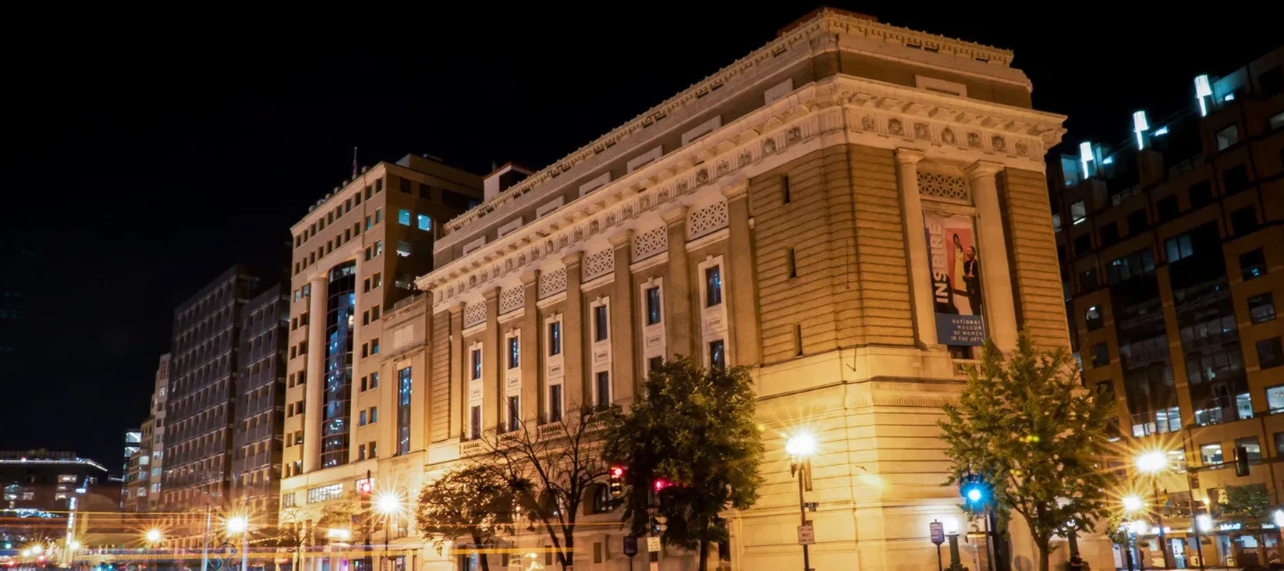 View of the National Museum of Women in the Arts building from outside showing the Neoclassical building from one corner. The building is a tan-colored stone with an arched doorway, long vertical windows, and detailed molding around the roof. It is dark outside, and street lamps shine brightly to illuminate the building.