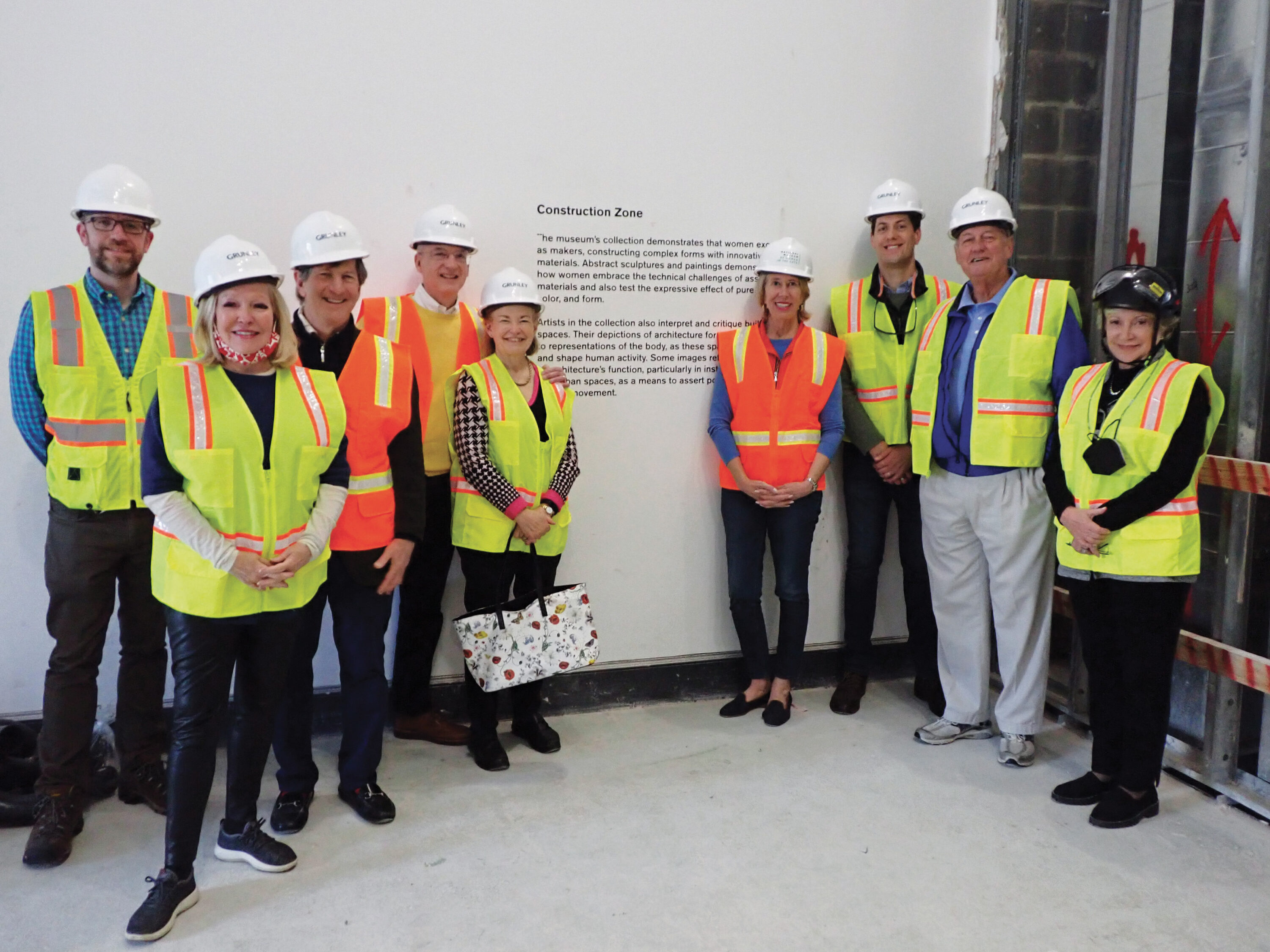 Two groups of four and five people in safety vests and helmets stand before a white wall with text on it. The men and women have a light skin tone and are adults of all ages. 