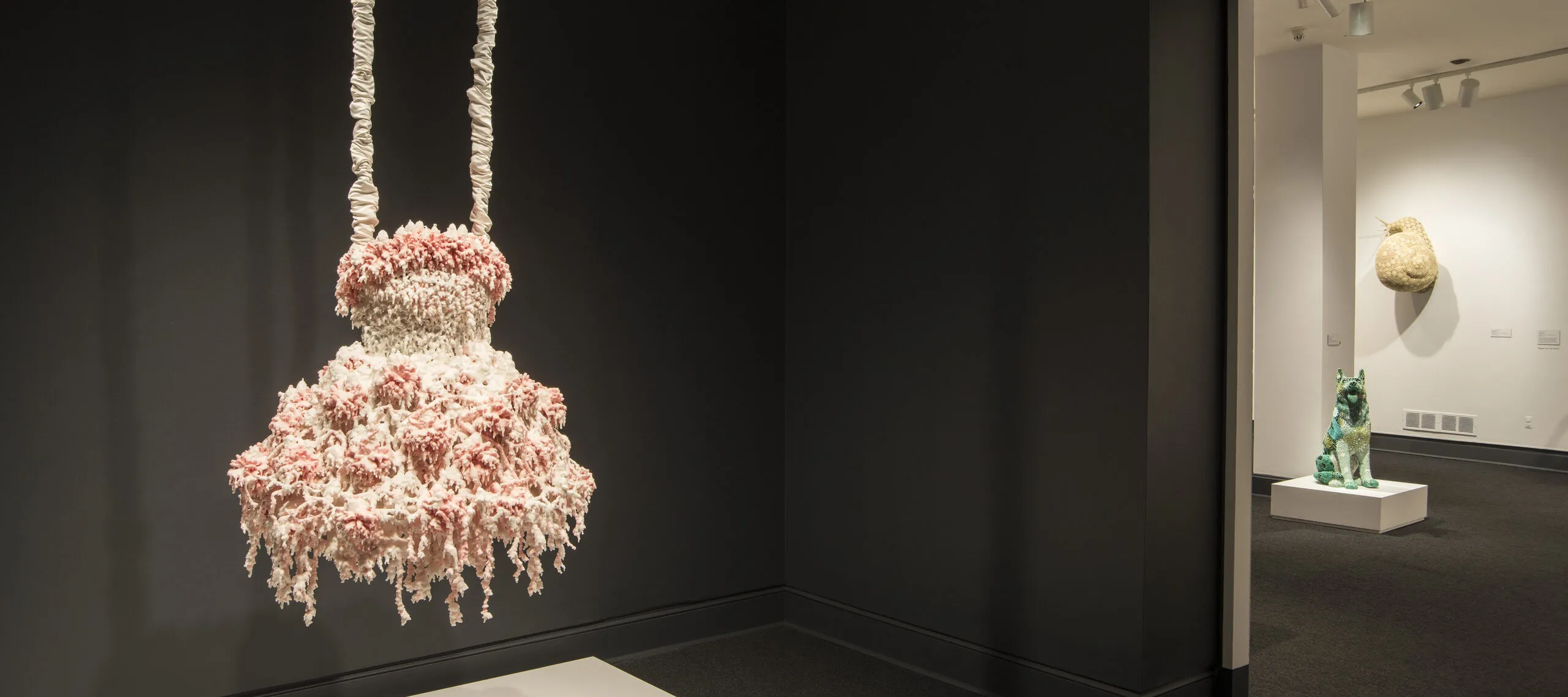 A sculpture hangs in a dark gallery. The sculpture comprises layers of melted pink and white wax that form a dress-like shape hanging from satin-wrapped chains. Its color, shape, and bumpy, lacy texture, evoke a frilly tutu, lavishly frosted wedding cake, or coral.