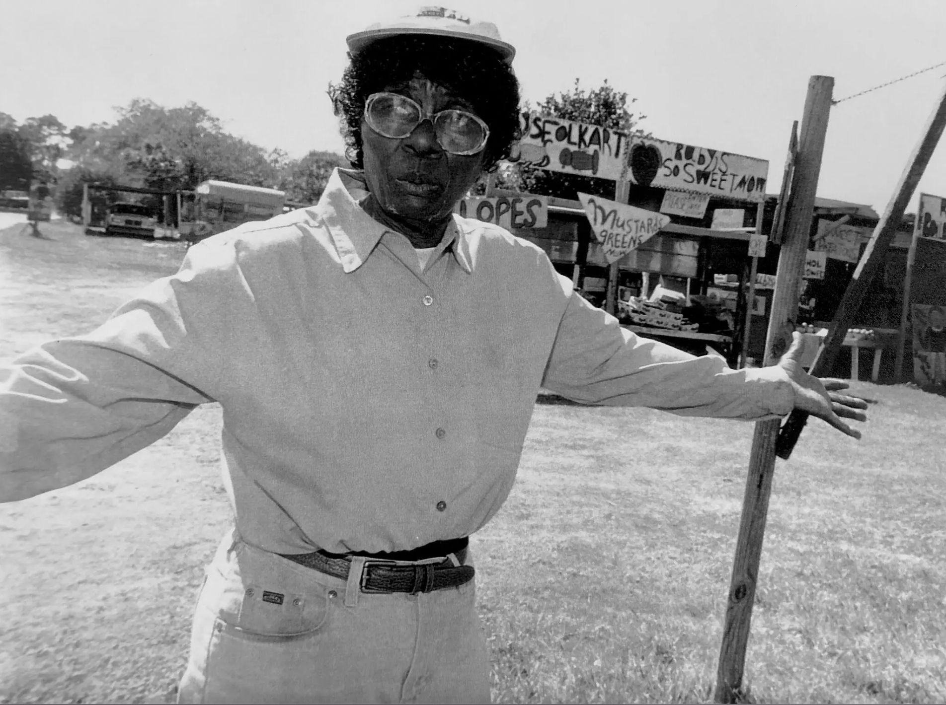 A black-and-white photograph of a woman with a dark skin tone wearing a white shirt, white jeans, and a white baseball hat. She is holding up her arms and slightly looks to the left; standing before a fruit stand with hand-painted signs. 