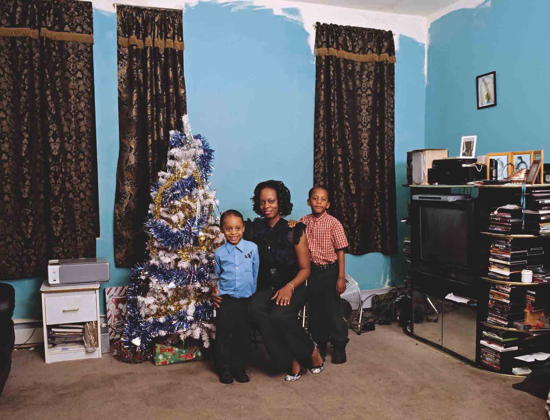 A woman and two boys with a medium-dark skin tone pose in front of a Christmas tree in formal attire. The Christmas tree is covered in gold, white, and blue tinsel. The light-blue wall behind them is only painted partly, giving the room an unfinished or impoverished air.