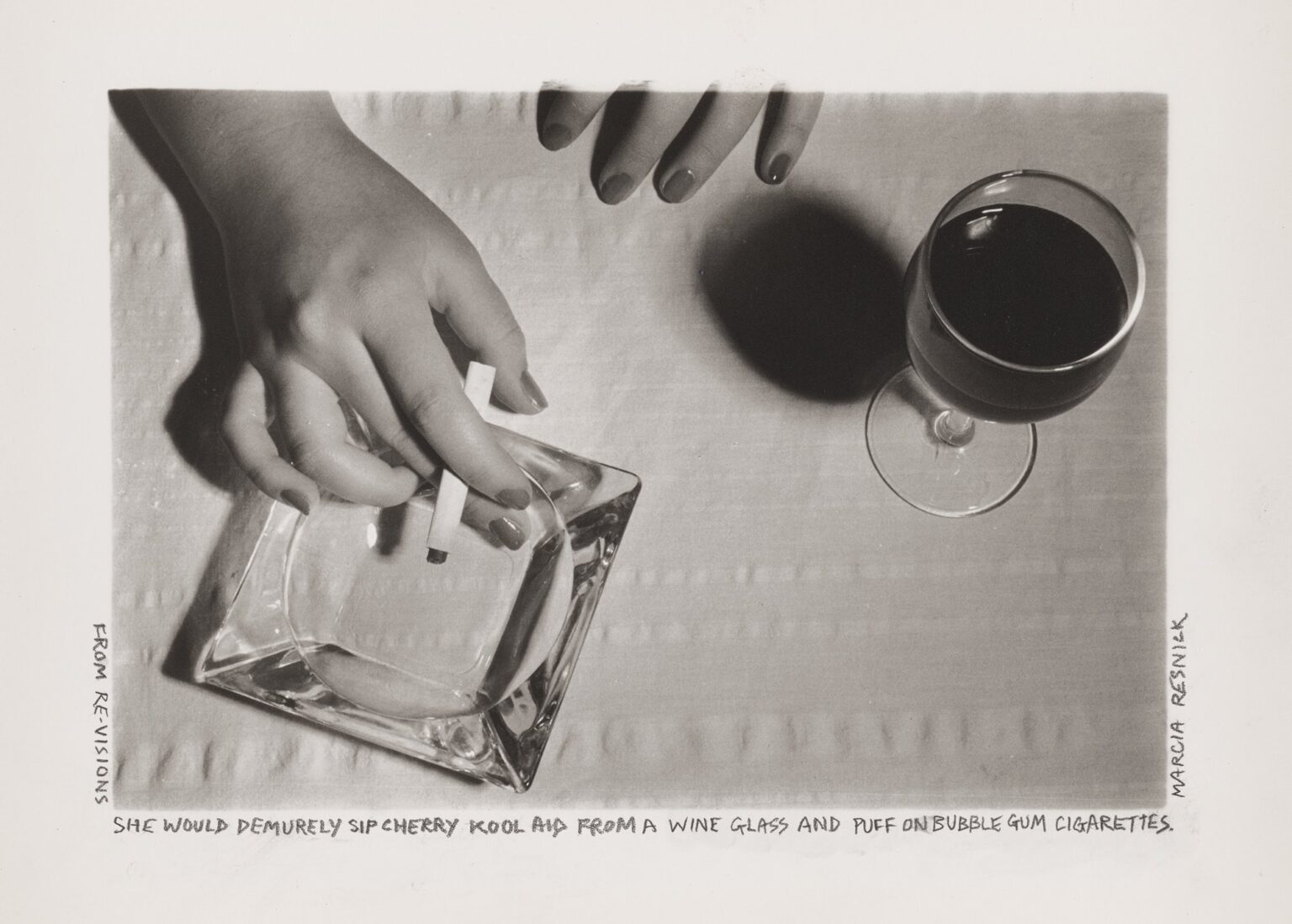 A black-and-white photograph depicts two hands on a dinner table, seen from above. One is holding a cigarette above a glass ashtray, and one is resting next to a full wine glass. Underneath the photograph, the text “She Would Demurely Sip Cherry Kool Aid From a Wine Glass and Puff on Bubble Gum Cigarettes” is written in pencil.