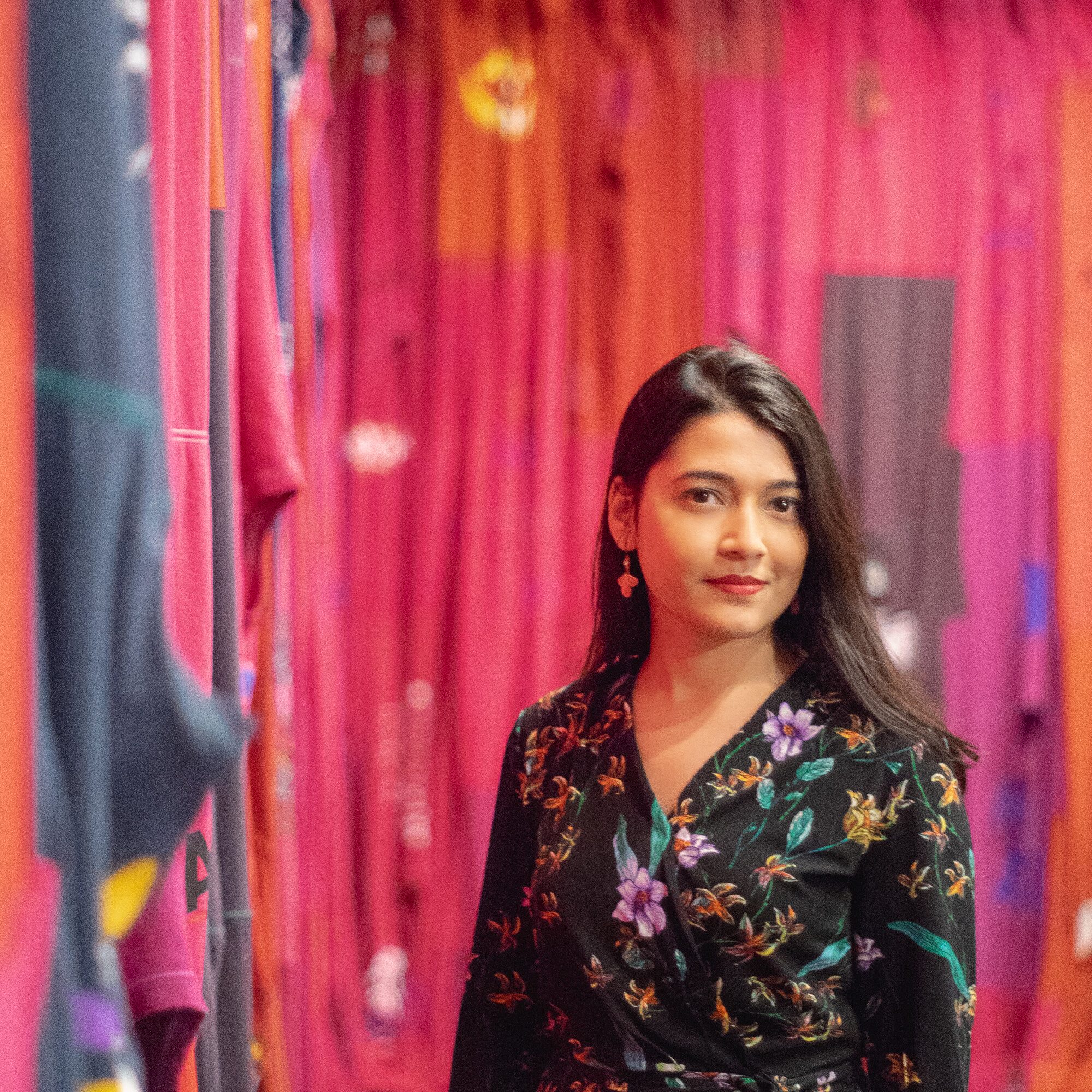 A photograph shows a woman with a medium skin tone and long black hair wearing a black blouse with a colorful floral print. Her head is slightly tilted to the right and she is smiling softly. In the background is a wall covered in pink and red textiles.