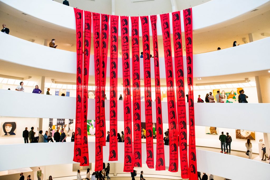 Several red banners with the face of a woman printed repeatedly on them and writing that says “WOMAN LIFE FREEDOM” hang from the ceiling of a rotunda in a museum. The bright red of the banners contrasts the white structure of the different levels within the museum. 