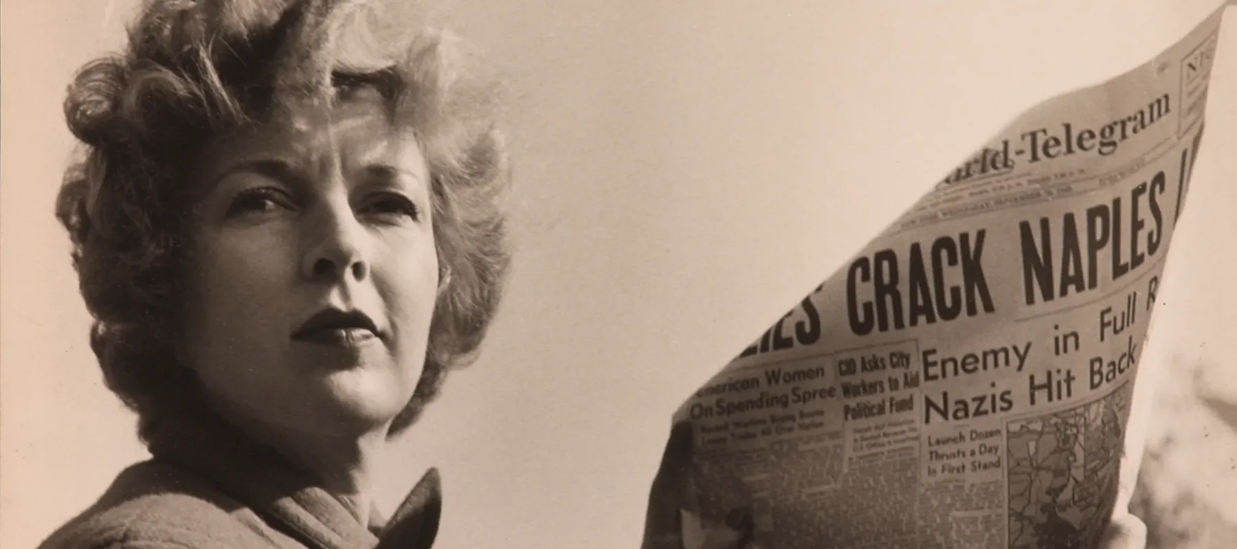 A black-and-white photograph of a light-skinned adult woman holding a newspaper with news about World War II. She wears a coat and her short, curly hair is caught in the wind.