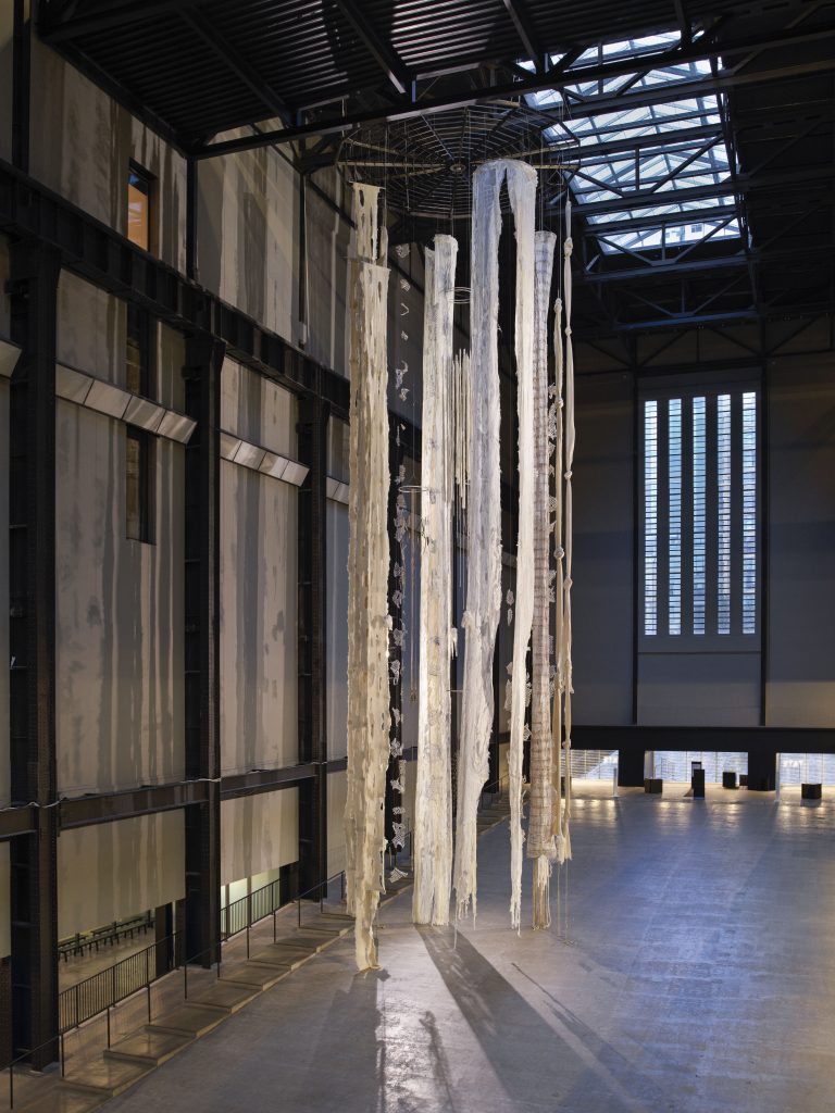 Long, white and beige textile strips hang from a ceiling of a vast industrial hall. The concrete and metal juxtapose the softness of the textiles that are attached to a round metal object. The sculpture looks similar like pieces of fabric attached to a fan, evoking a breezy and airy feeling in the industrial surrounding.