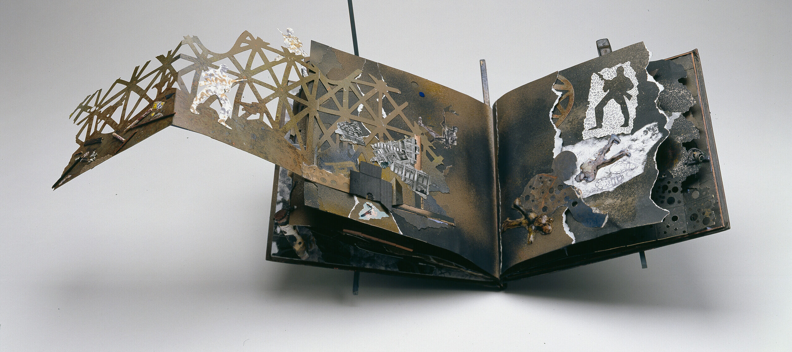 An artist book sits open on a grey background. The open pages reveal a collage filled with scenes of war: soldiers, barbed wire, and ammunition. A toy version of a military helicopter protrudes from the cover of the book.