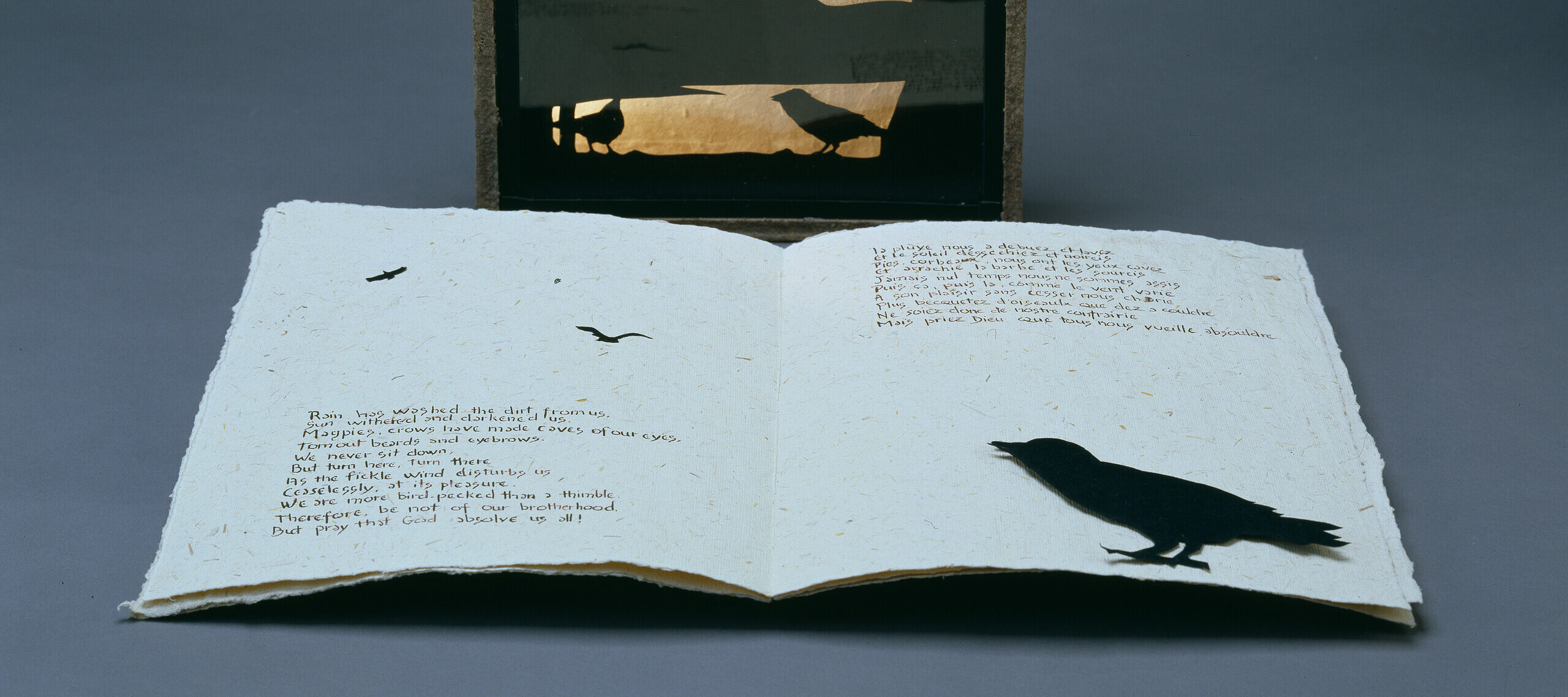 A book is opened to a page with black outlines of crows and a paragraph is written on each page in neat printed handwriting. Standing behind the book, a stone frame outlines the shadow of three people hanging from ropes with crows surrounding their bodies.