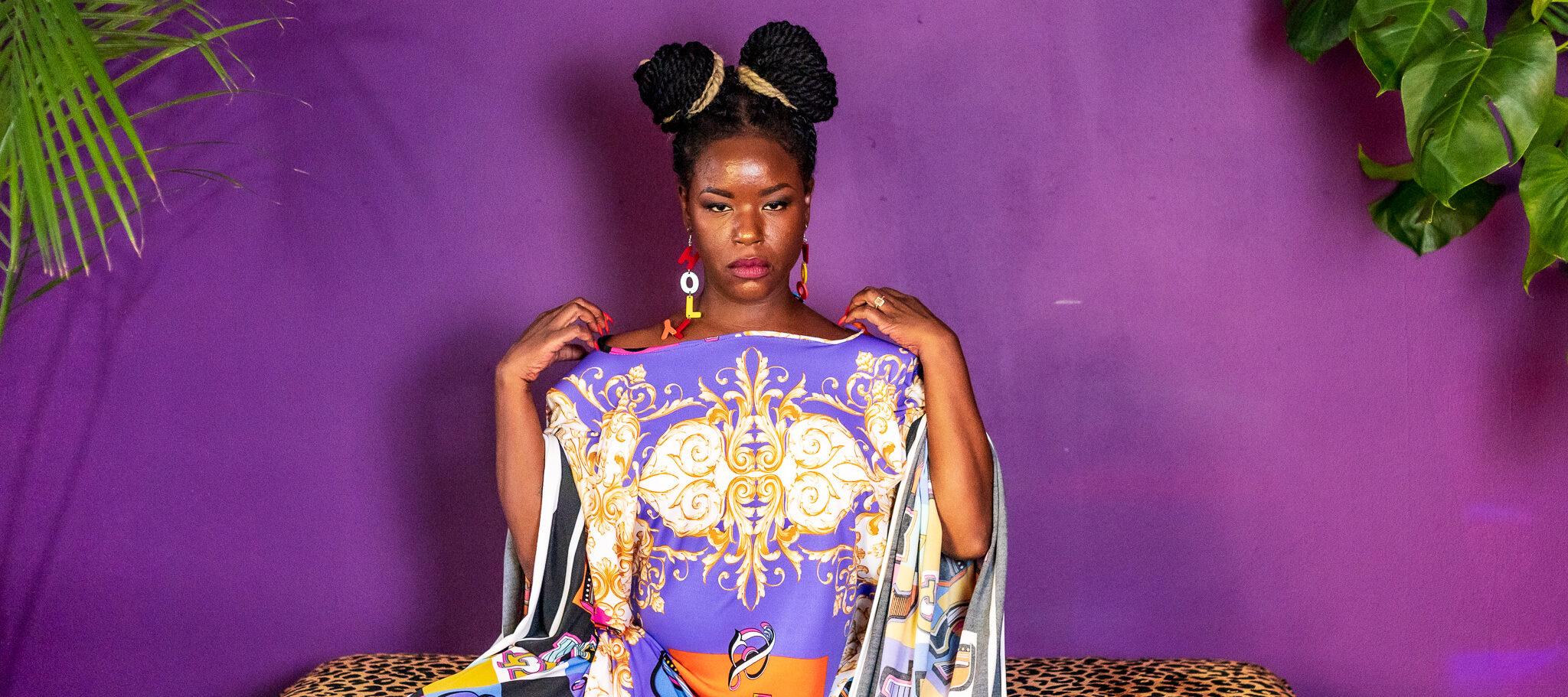 A medium-dark skinned person in a purple, white, and gold dress sits on an cheetah print bench in front of a purple wall. They stare directly in front of them with their hands on their shoulders.
