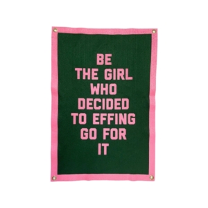 A wool rectangular banner with pink, all caps lettering that says "Be the girl who decided to effing go for it" on a dark green background. 
