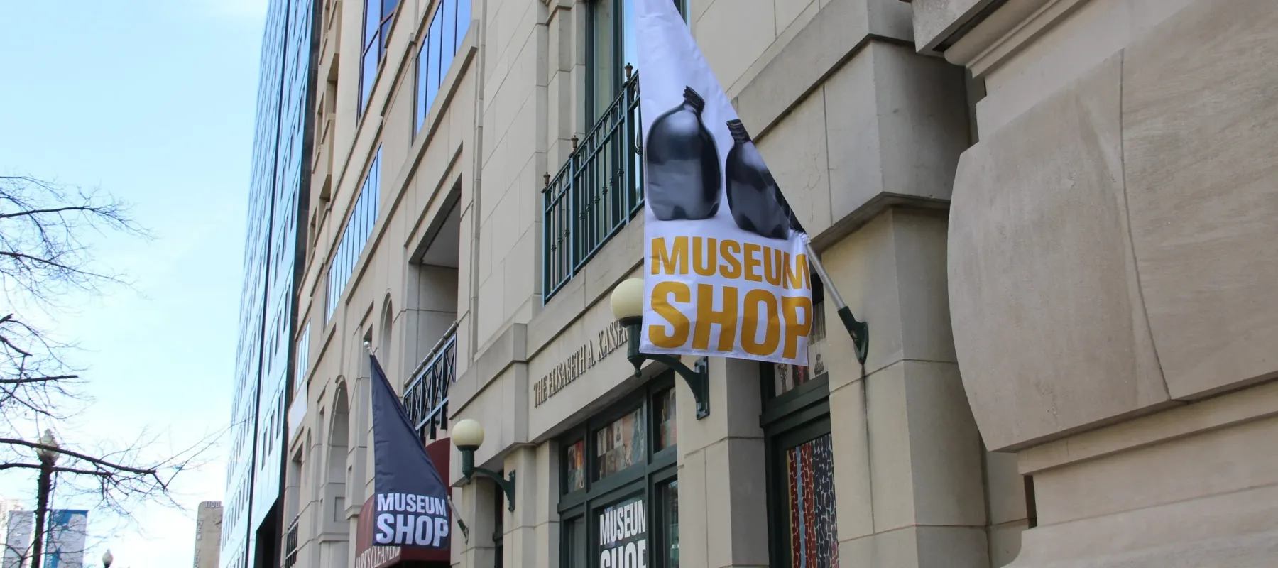 A white flag hangs on the facade of a stone building. The flag has an image of two black ceramic vases and "Museum Shop" written in gold letters below them.