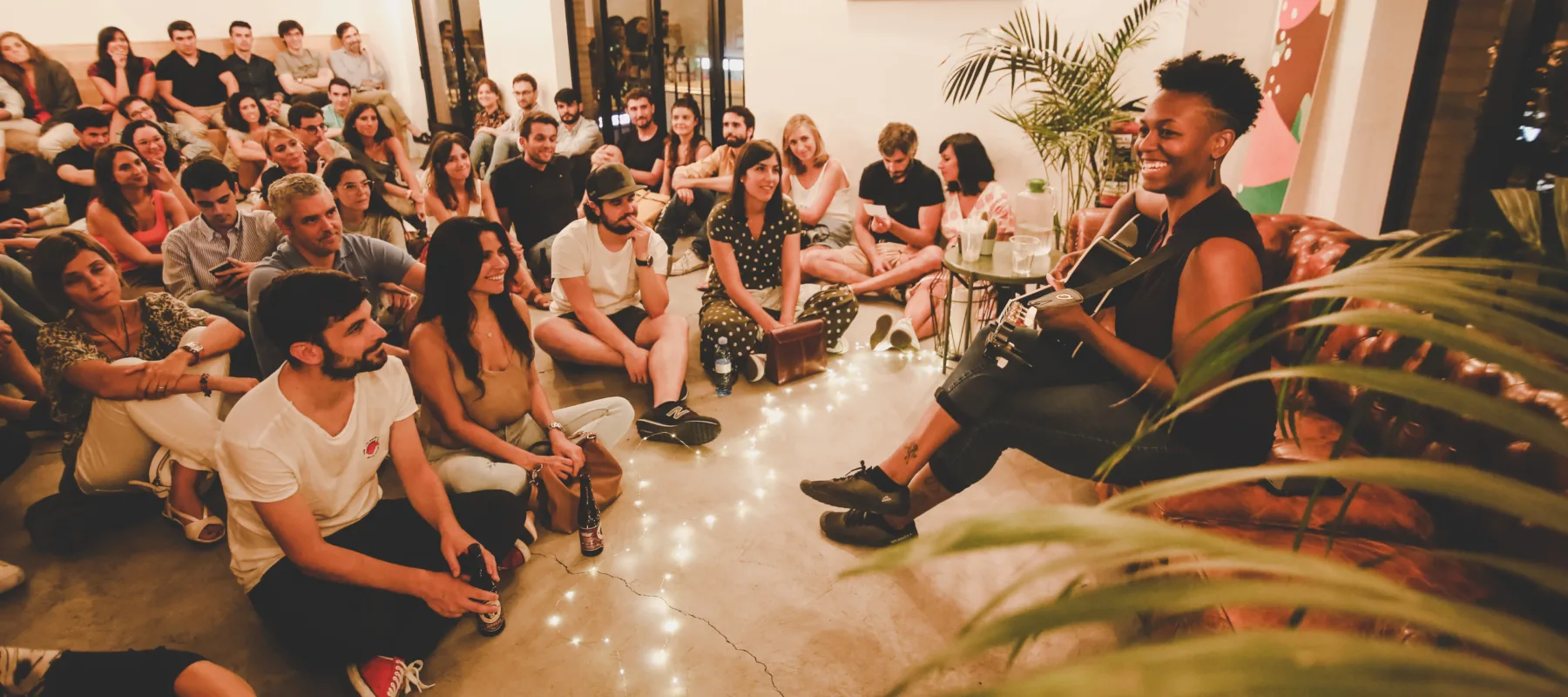 A medium-skinned woman with short curly hair sits on a leather couch playing a guitar. She is surrounded by an audience sitting on the floor watching her performance.