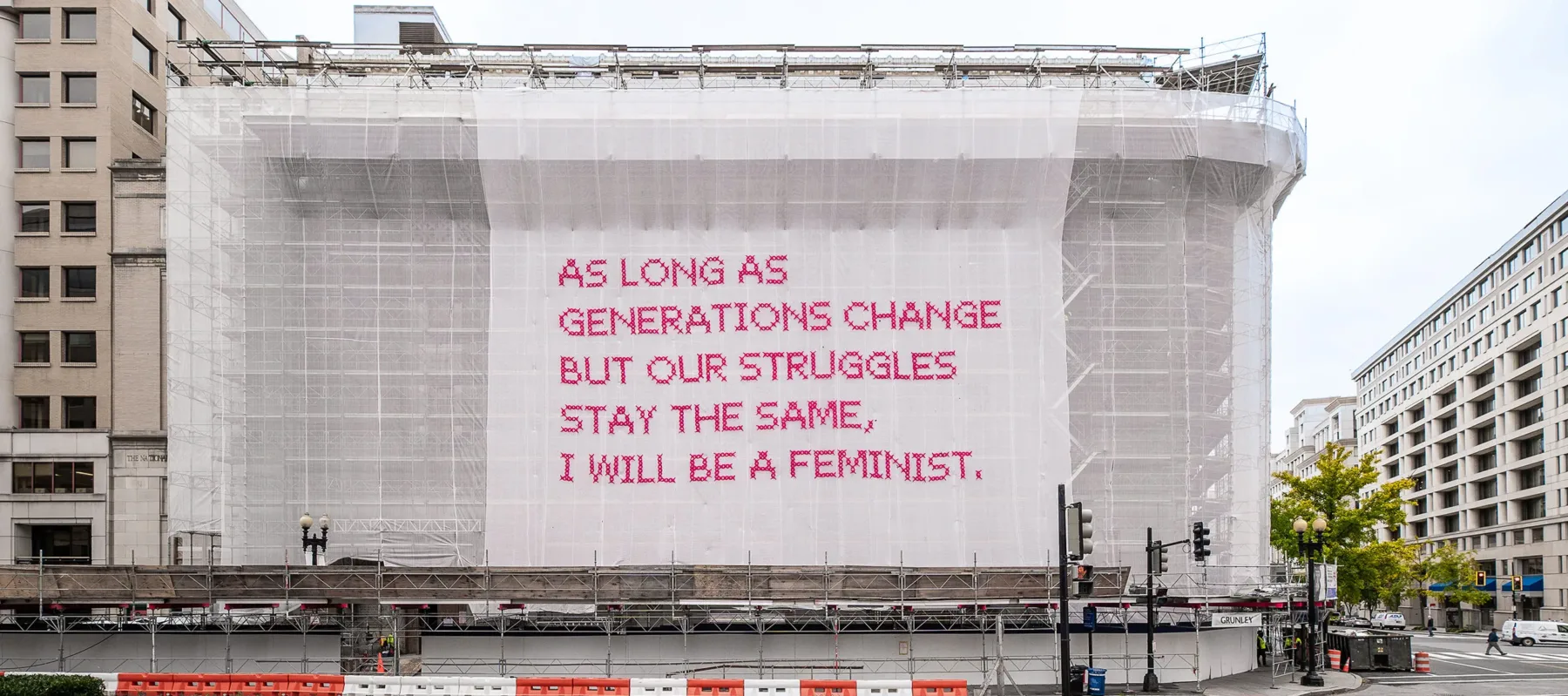 White gauzy fabric hangs in front of scaffolding surrounding a city building. Bold magenta letters on the fabric read, “As long as generations change but our struggles stay the same, I will be a feminist.”