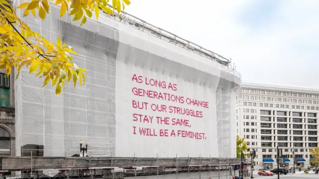 White gauzy fabric hangs in front of scaffolding surrounding a city building. Bold magenta letters on the fabric read, “As long as generations change but our struggles stay the same, I will be a feminist.”