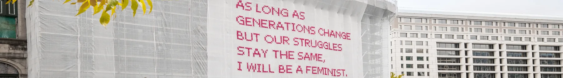 White gauzy fabric hangs in front of scaffolding surrounding a city building. Bold magenta letters on the fabric read, “As long as generations change but our struggles stay the same, I will be a feminist.”