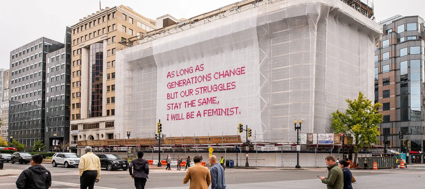 White gauzy fabric hangs in front of scaffolding surrounding a city building. Bold magenta letters on the fabric read, “As long as generations change but our struggles stay the same, I will be a feminist.” Several passersby cross the street in front of the building.