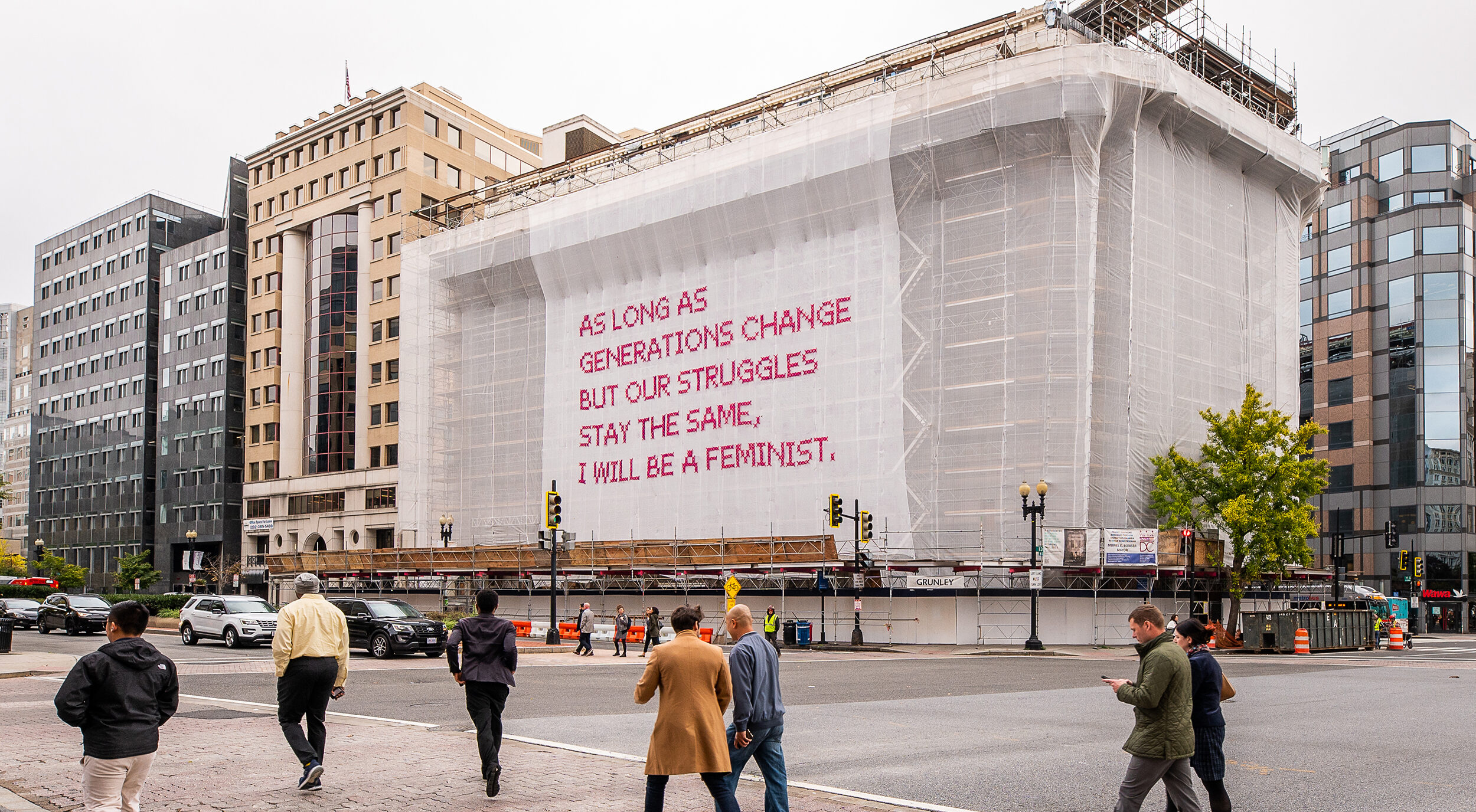 White gauzy fabric hangs in front of scaffolding surrounding a city building. Bold magenta letters on the fabric read, “As long as generations change but our struggles stay the same, I will be a feminist.” Several passersby cross the street in front of the building.