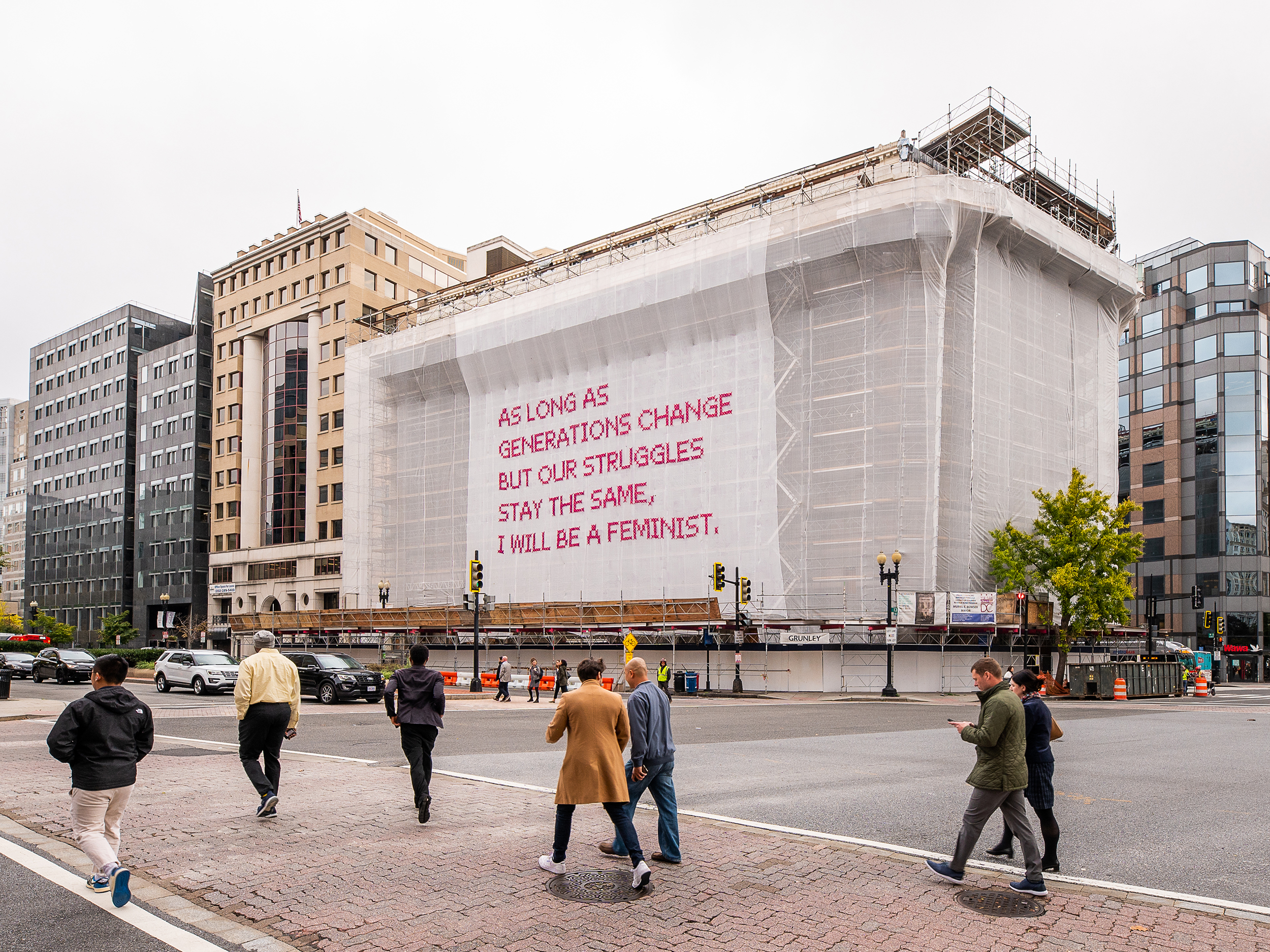White gauzy fabric hangs in front of scaffolding surrounding a city building. Bold magenta letters on the fabric read, “As long as generations change but our struggles stay the same, I will be a feminist.” Several passersby cross the street in front of the building.