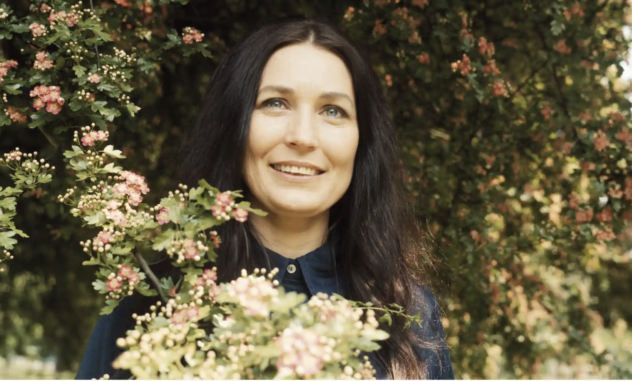 A woman with a light skin tone and long dark hair wears a dark-blue Western button-up shirt amid a cherry blossom tree. She is gazing into the distance and smiling.