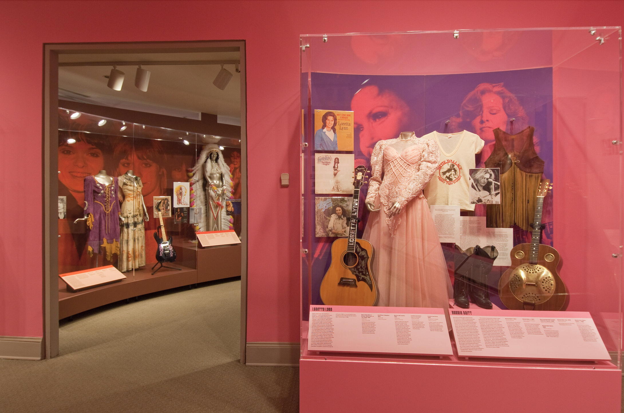 A view into a gallery space with pink walls and glass cases including guitars, women’s country western outfits, and records picturing women.